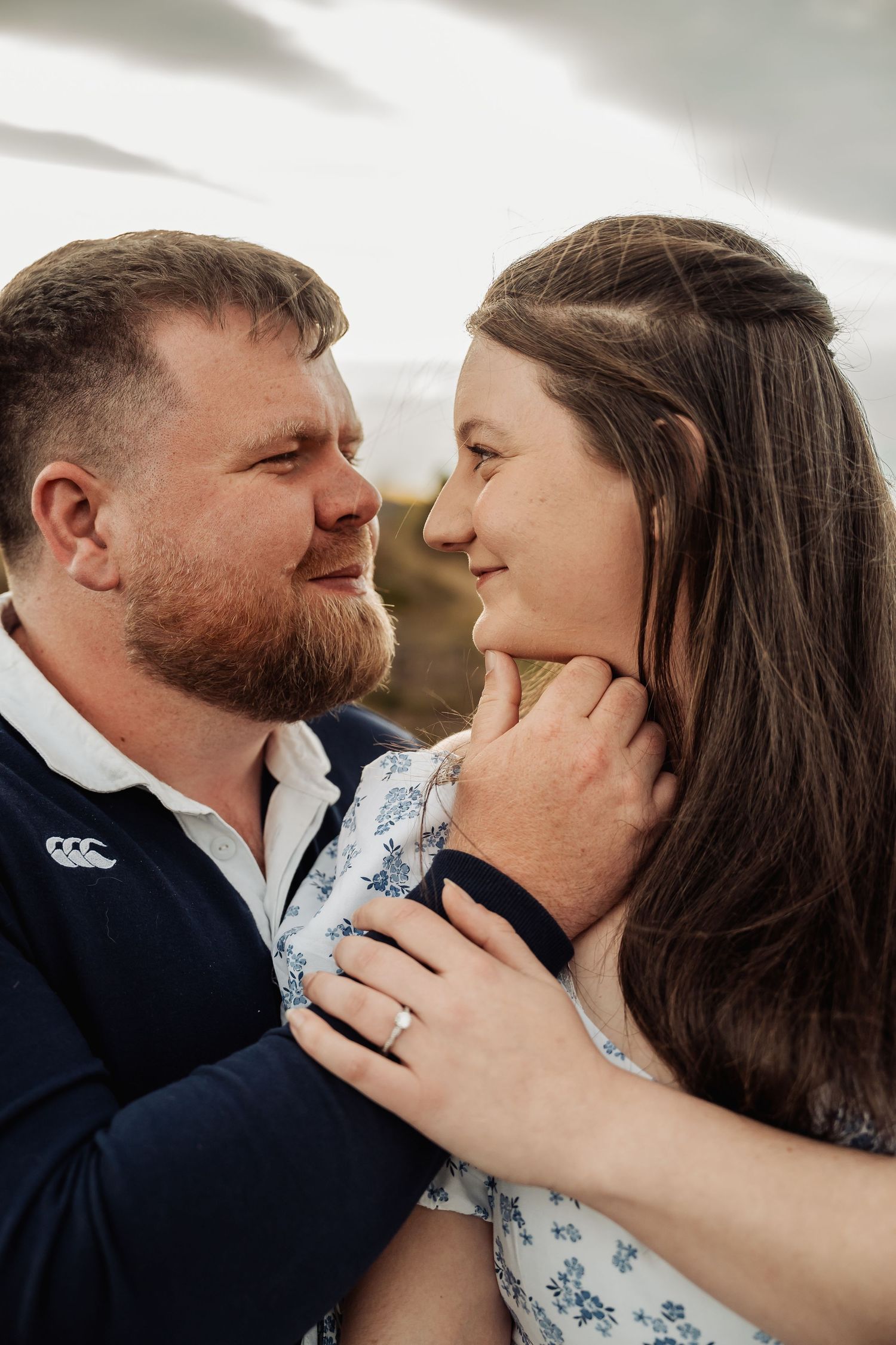 A couple gazes lovingly at each other outdoors, the woman wearing an engagement ring as the man gently holds her face.
