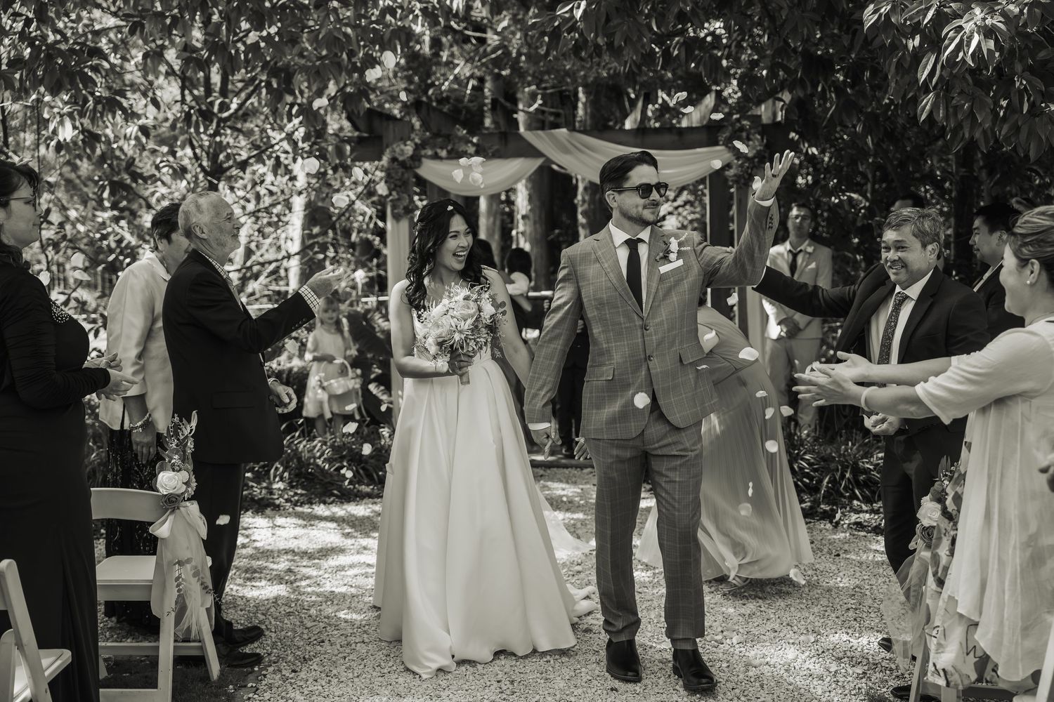 Black and white photo of newlyweds walking back down the aisle as guests celebrate with confetti at outdoor wedding.