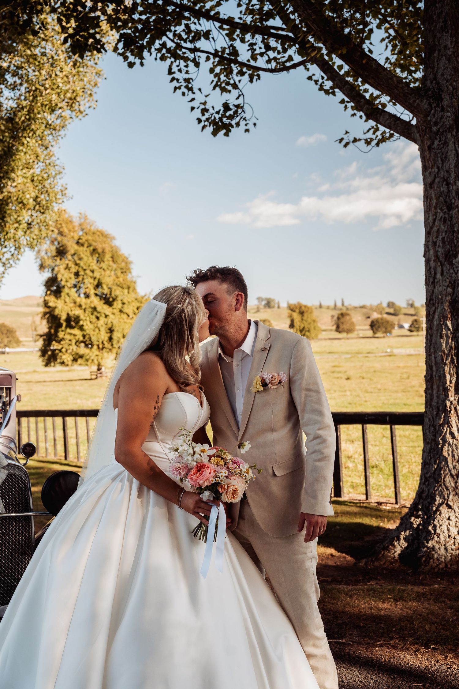 Bride and groom share a kiss under a tree with scenic countryside views on their wedding day.