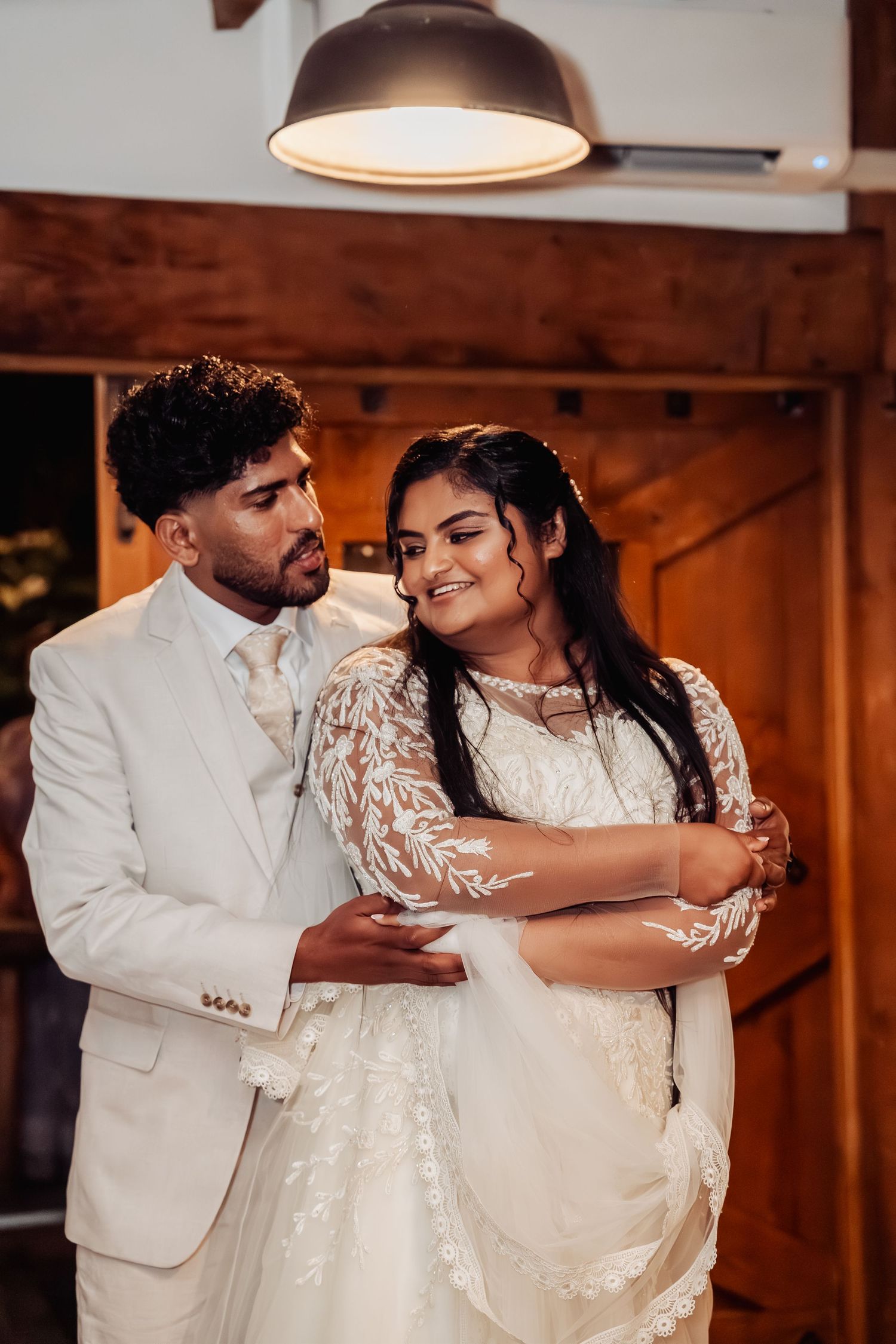 Bride in lace gown and groom in white suit share a warm embrace indoors under warm lighting.