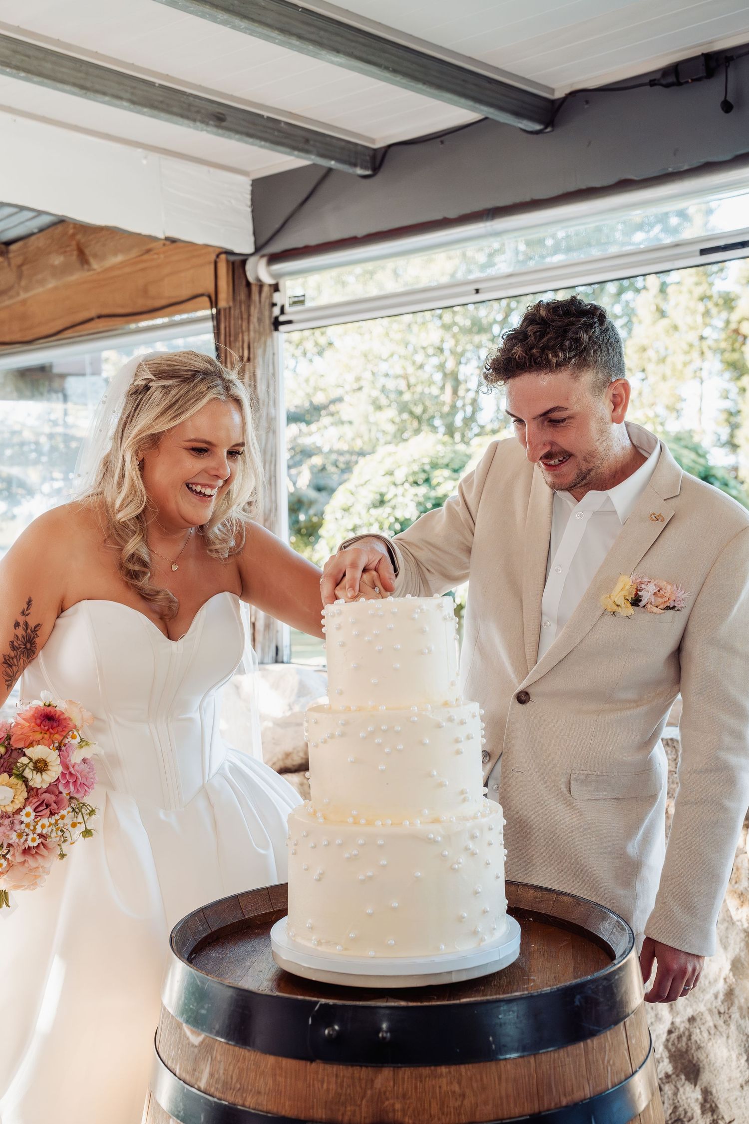 Smiling bride and groom cut their elegant three-tier white wedding cake on a barrel together.