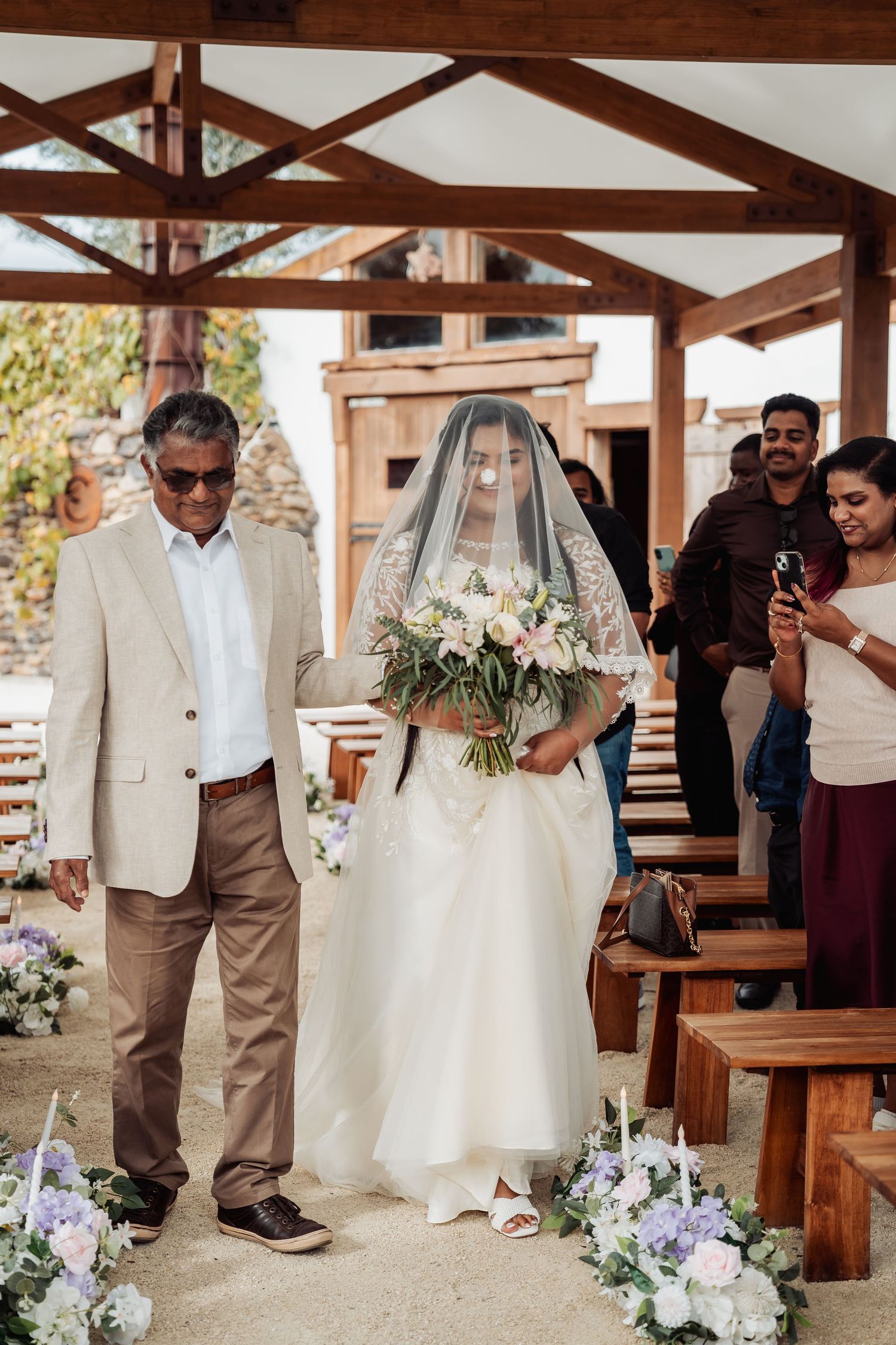 Bride walking down aisle with father in rustic outdoor wedding chapel with floral decorations.