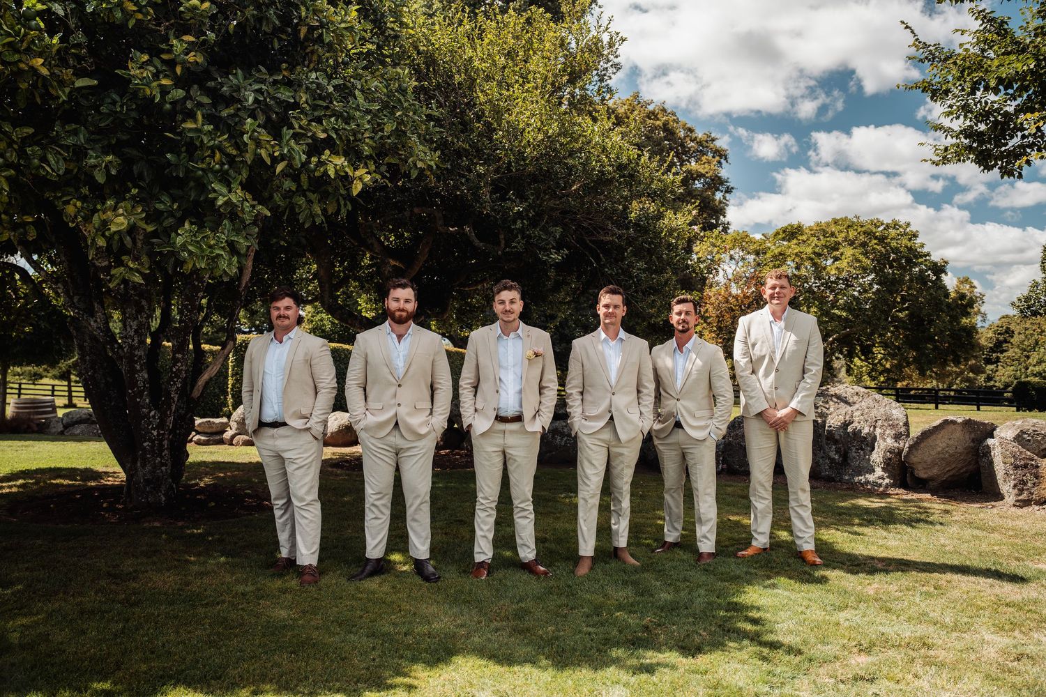 Groom and groomsmen posing together in matching beige suits on a sunny green lawn.