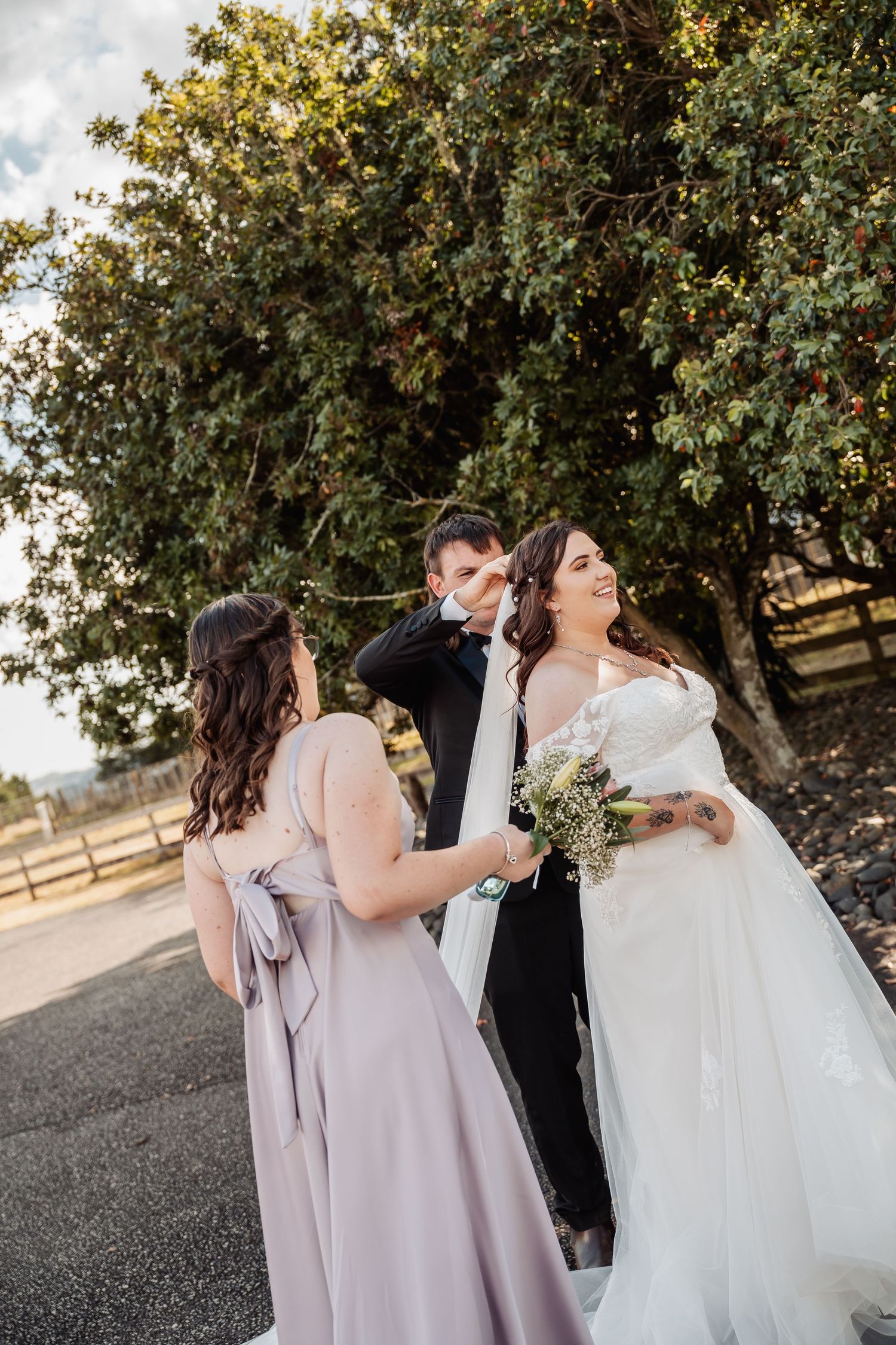 Bride in white gown holds bouquet while bridesmaid adjusts dress as groom wipes tears outdoors.