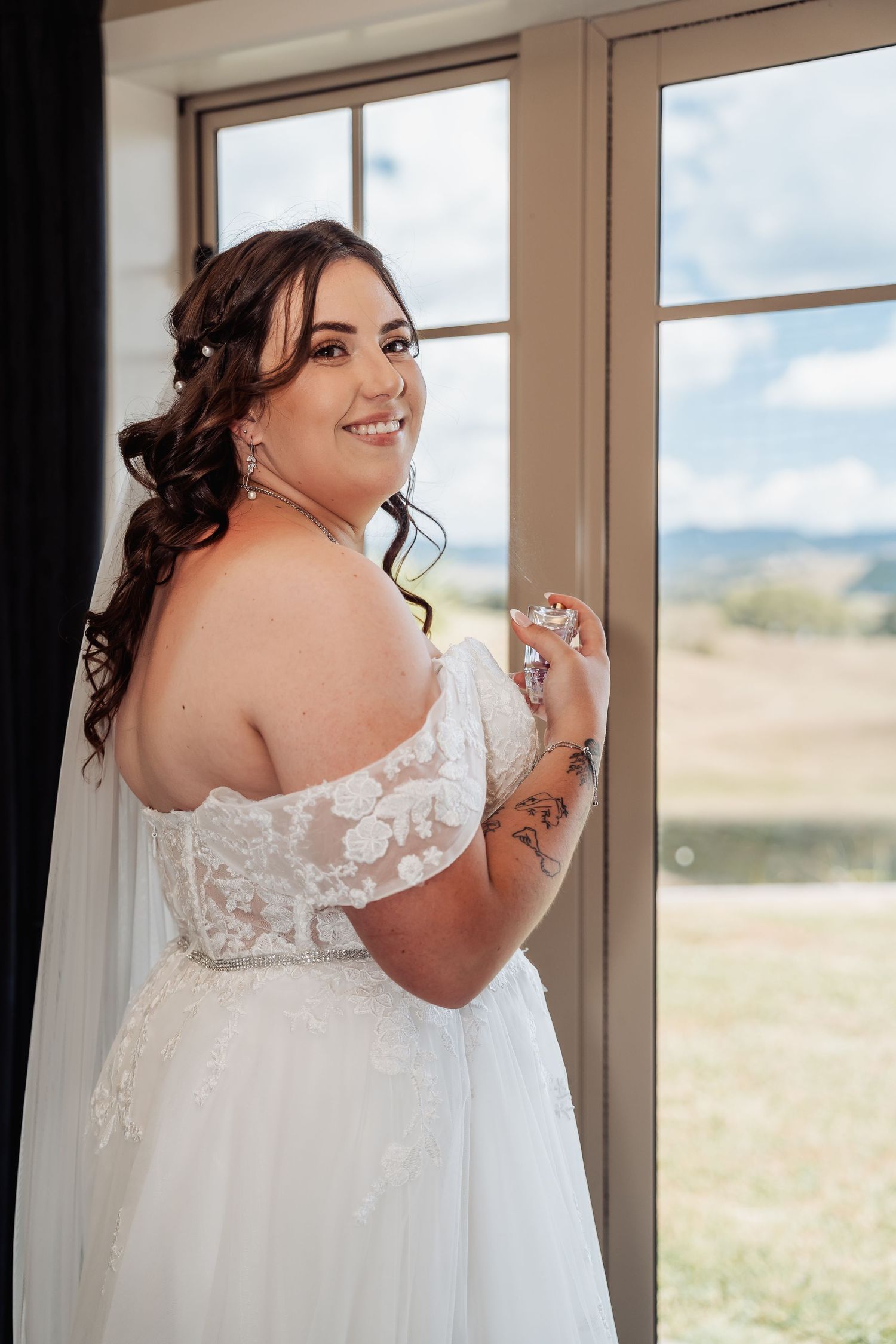 Smiling bride in off-shoulder lace wedding dress holds perfume bottle by window with scenic view.