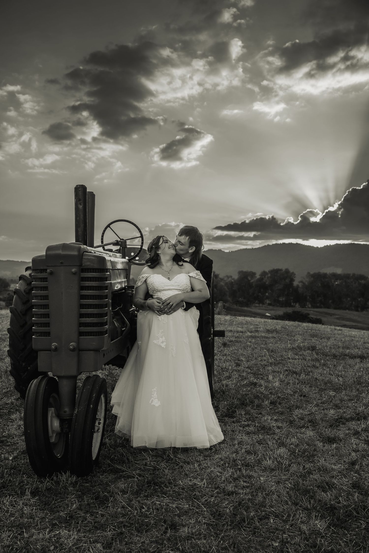 Black and white photo of couple kissing beside vintage tractor with dramatic sunset sky backdrop.