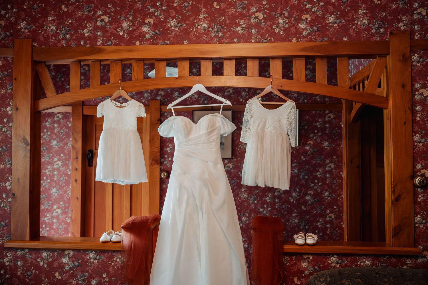 Wedding dress and two bridesmaid dresses hanging on wooden mirror frame with floral wallpaper.