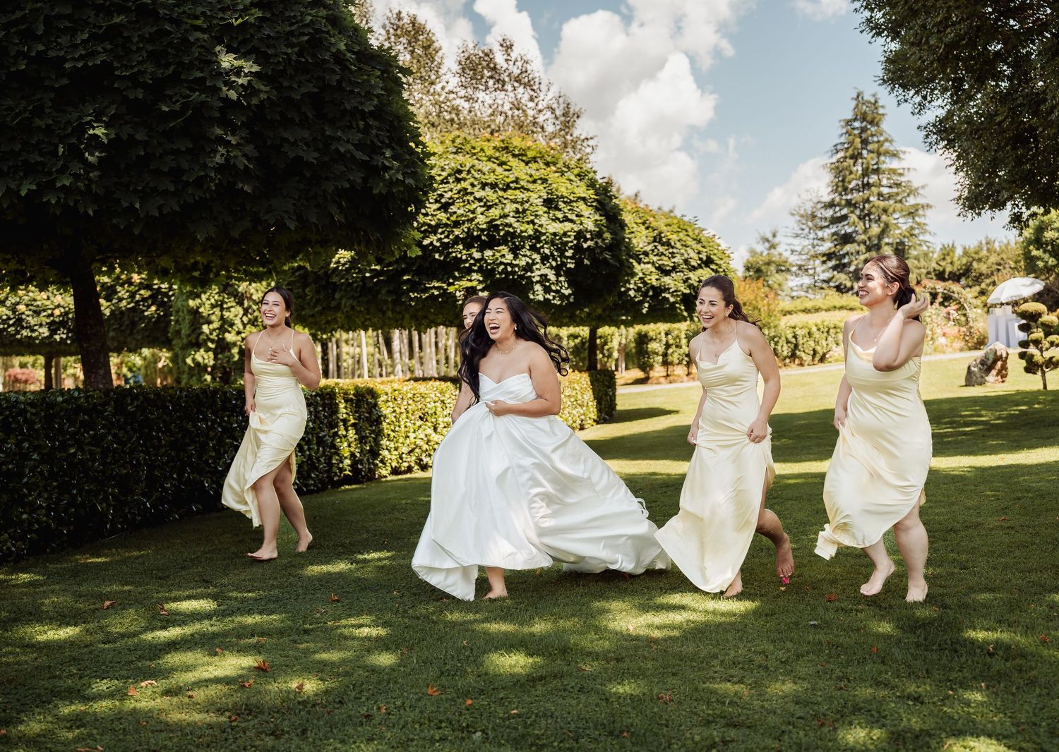 Bride and bridesmaids in white dresses laughing and running together on lush green lawn at outdoor wedding venue.