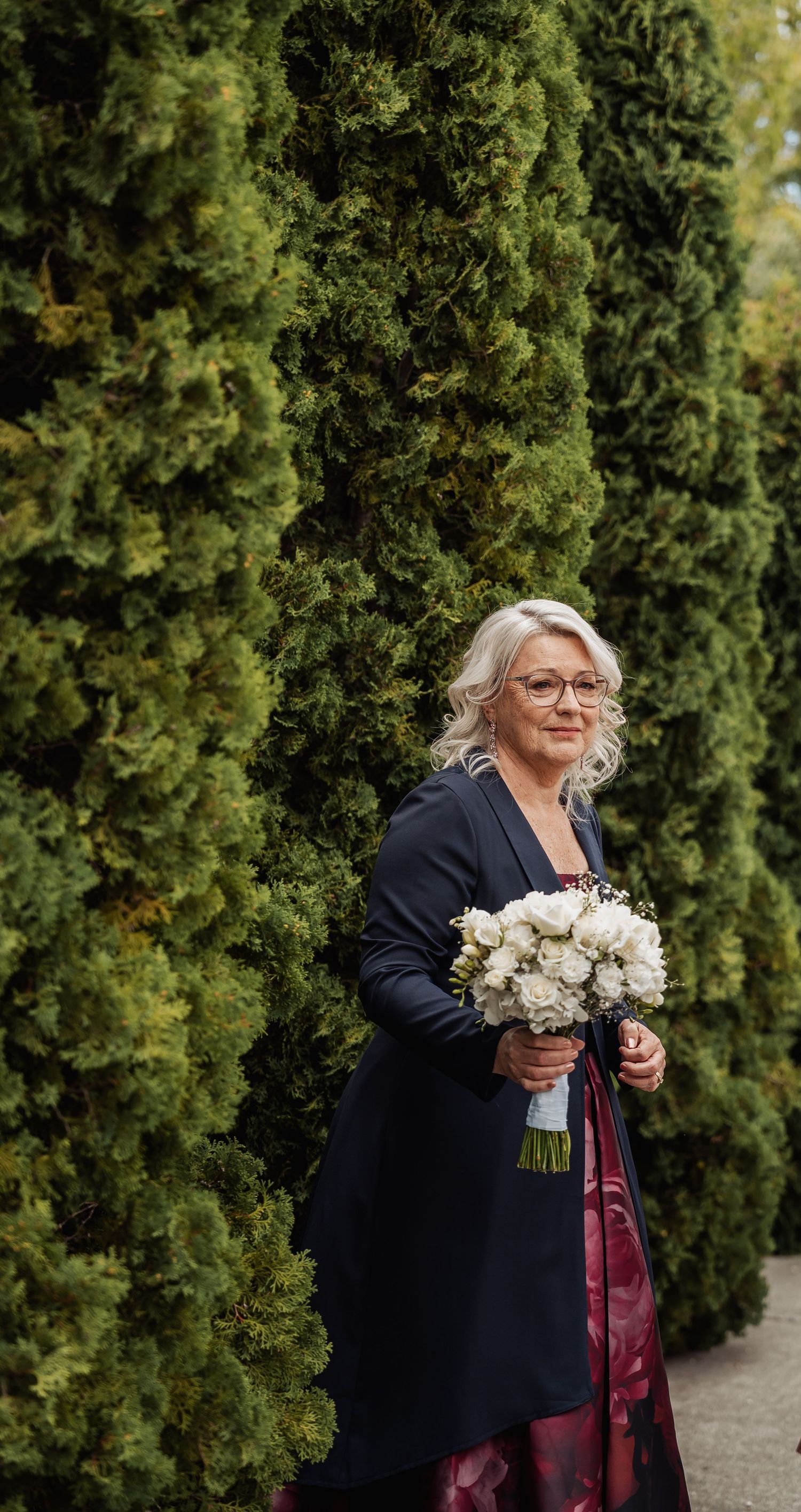 Elegant older woman in a navy blazer and burgundy dress holding a white rose bouquet beside tall green hedges.