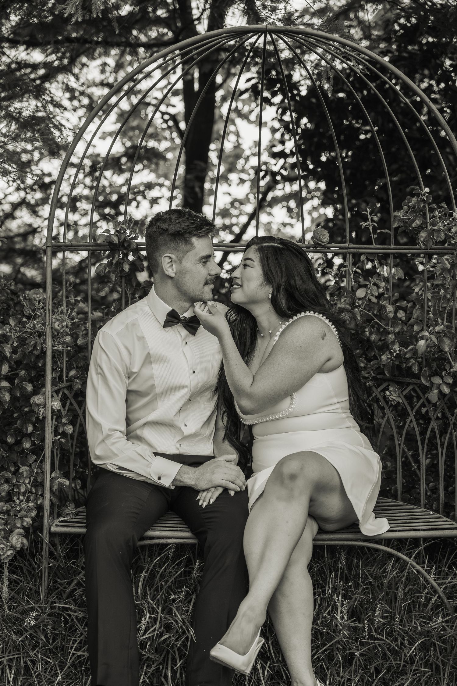 Black and white photo of newlyweds sitting together on ornate bench inside decorative garden gazebo.