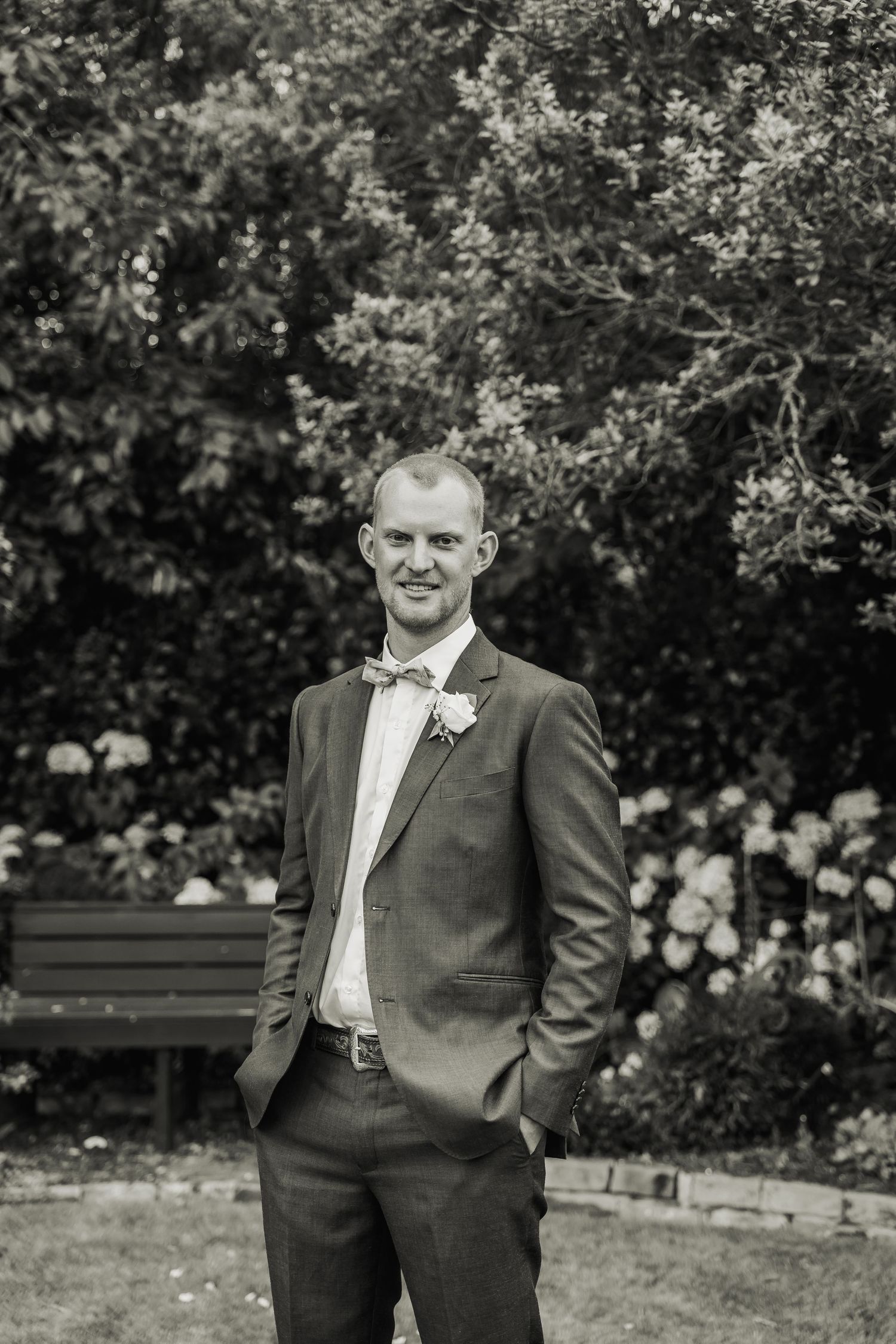 Black and white portrait of a smiling groom in a suit with bow tie and boutonniere in a garden.