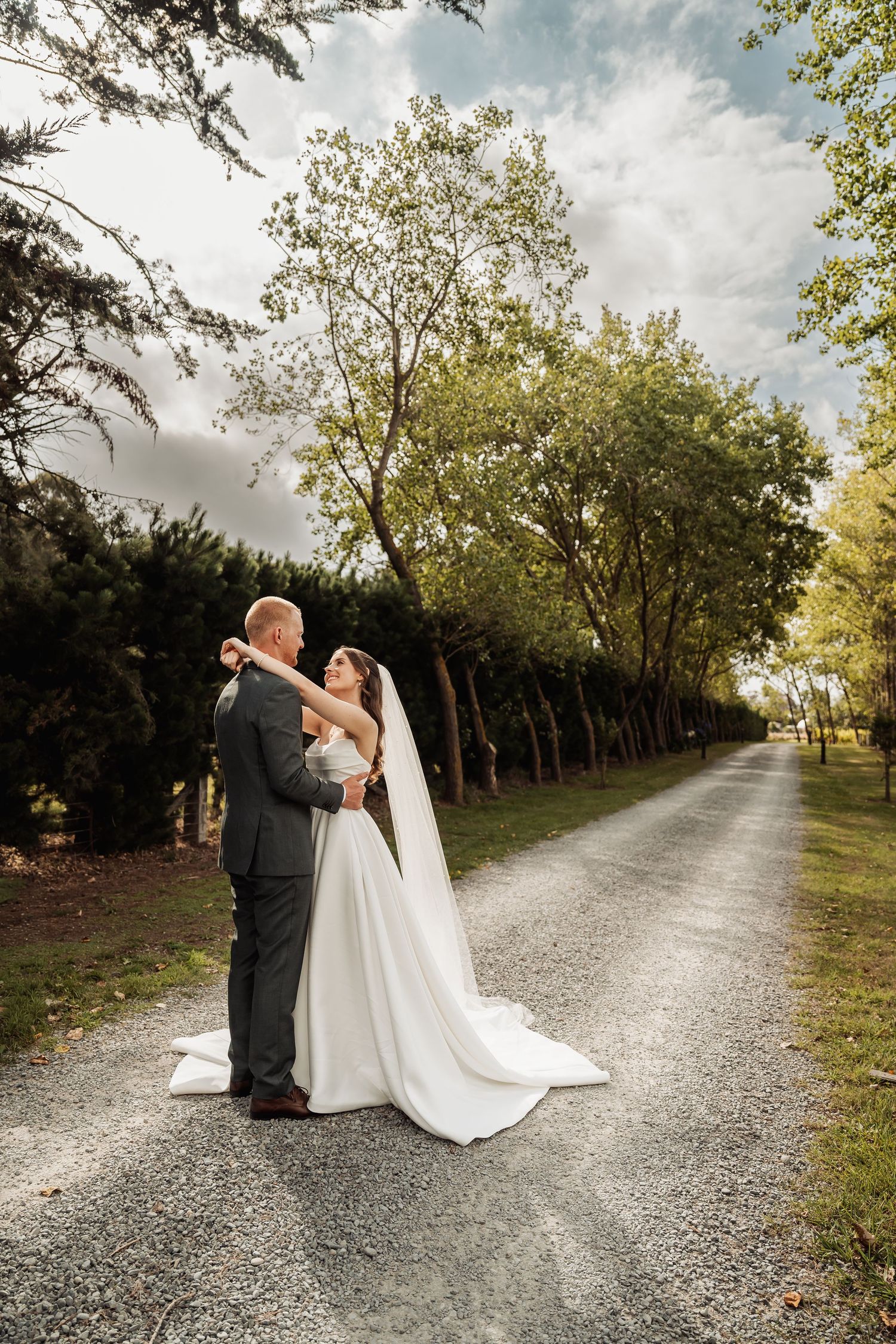 Bride and groom embrace on a tree-lined gravel path under a bright cloudy sky.