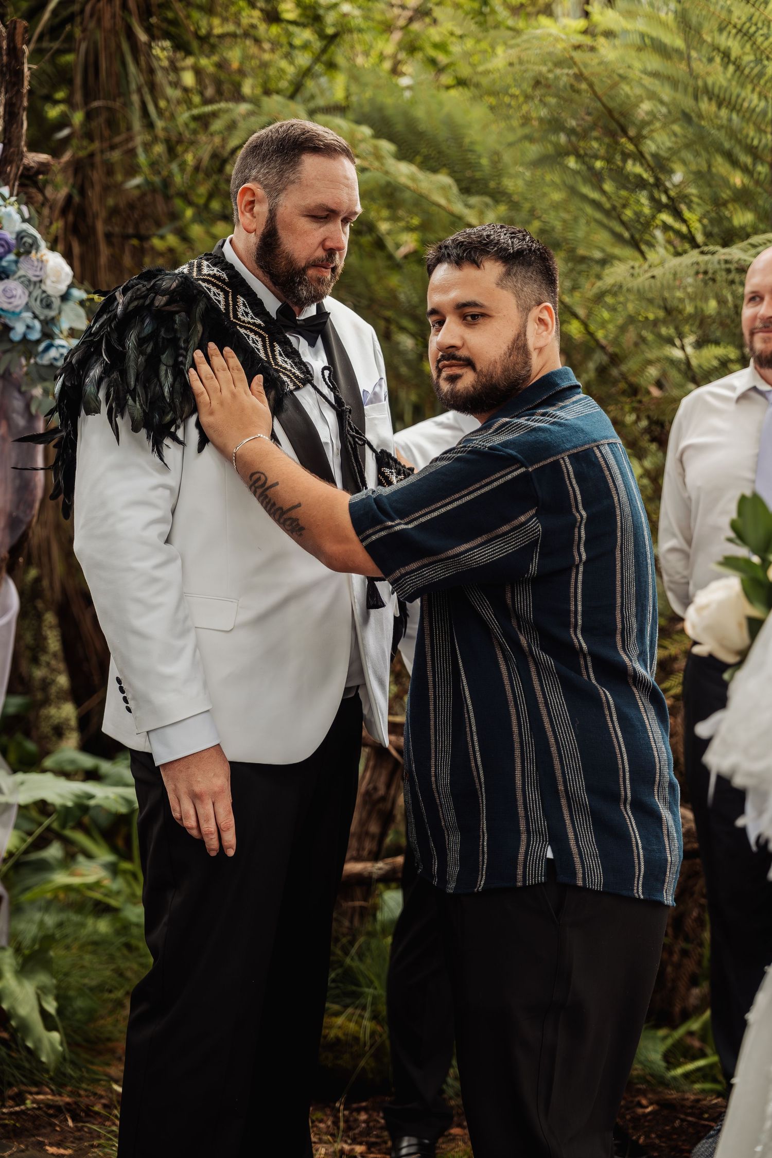 Man adjusting traditional Maori feather cloak on groom wearing white tuxedo at outdoor forest wedding.