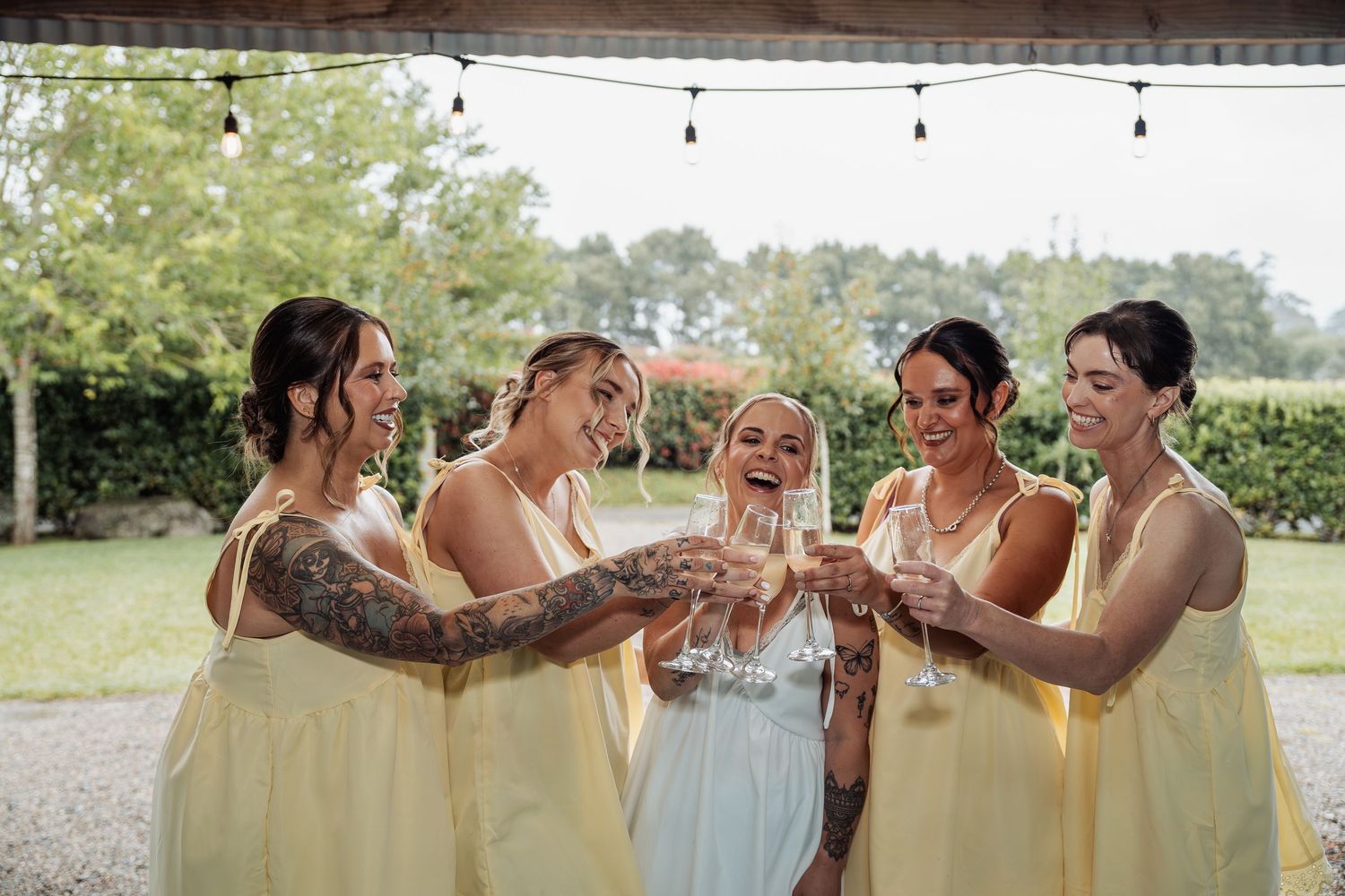 Bride in white dress toasting champagne with four bridesmaids in yellow dresses at an outdoor wedding celebration.