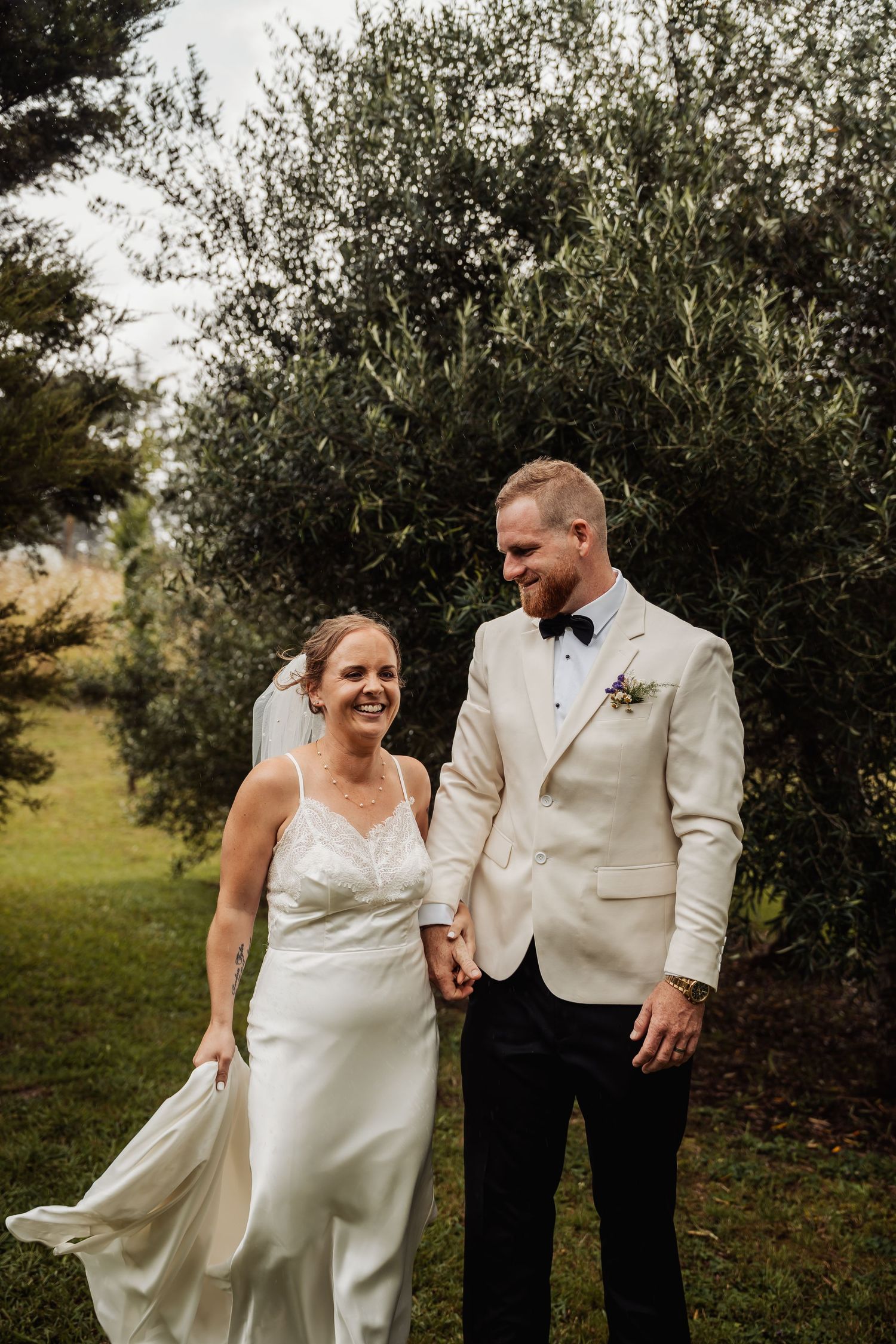 Bride and groom holding hands and laughing while walking through a lush green garden together.