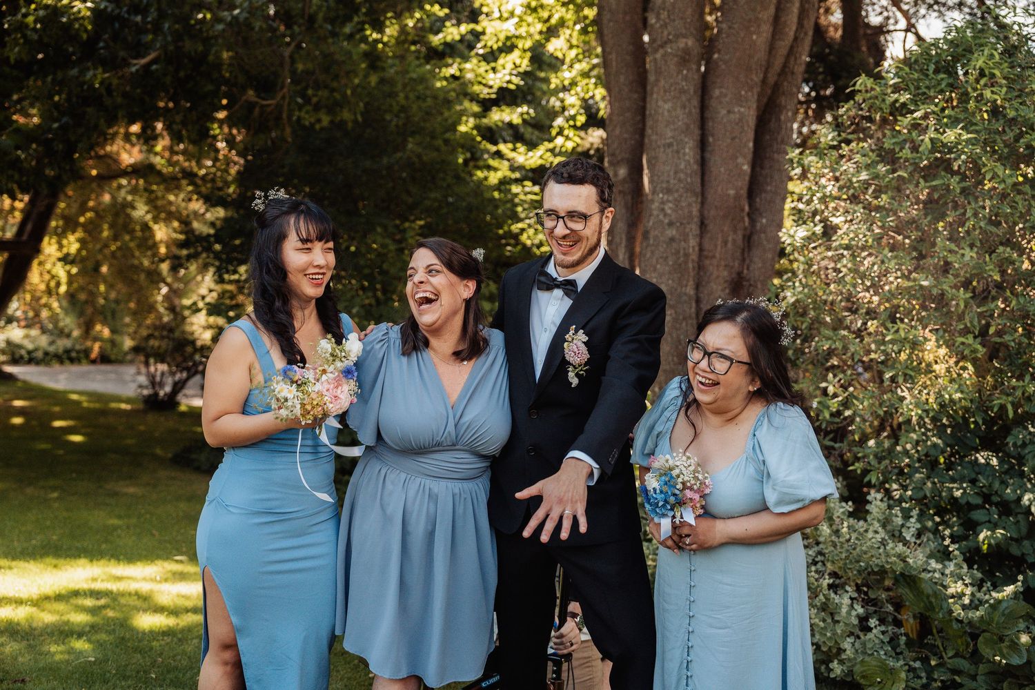 Groom in tuxedo laughs with three bridesmaids in blue dresses outdoors.