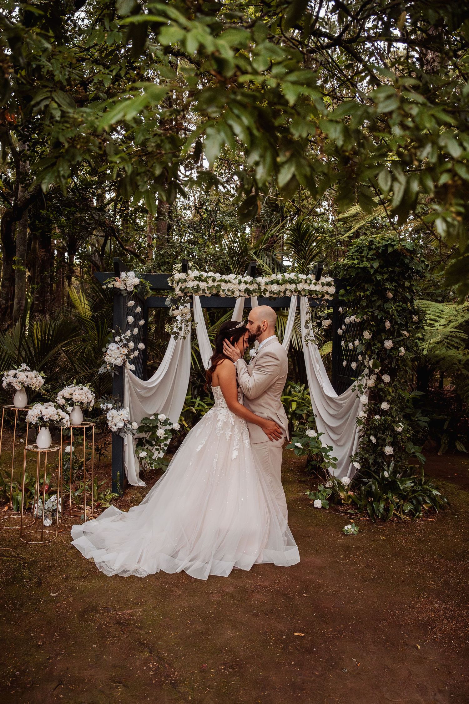 Bride and groom share romantic kiss under floral arch with white draping surrounded by lush greenery at outdoor wedding ceremony.