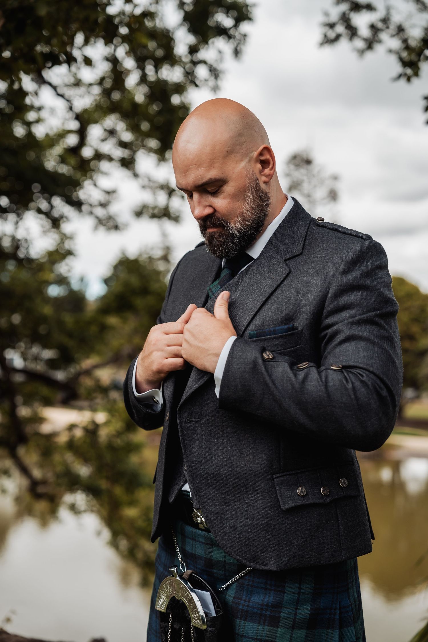 Bald bearded groom in Scottish kilt and tweed jacket adjusting his tie outdoors near a pond.