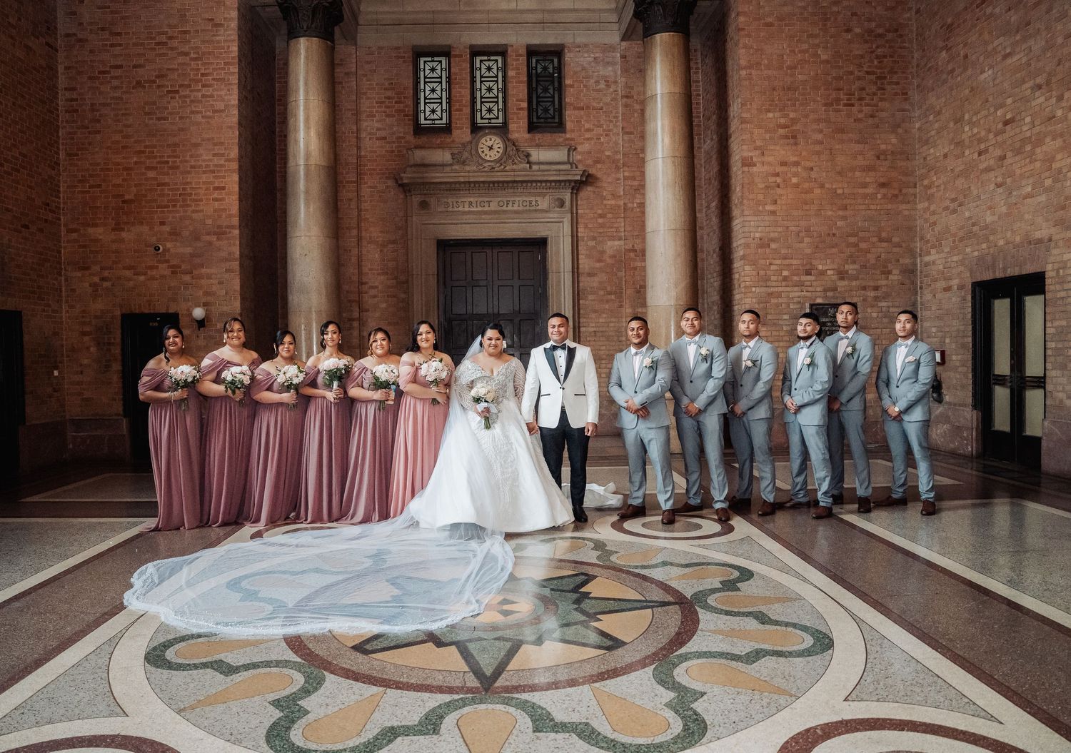 Wedding party in gray suits poses in front of a grand brick wall with ornate floor designs.