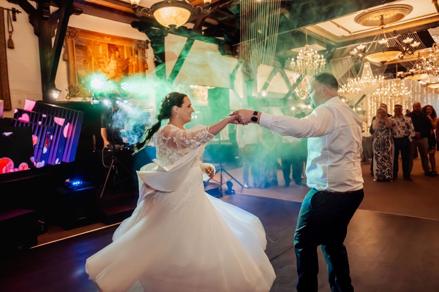 Wedding couple dances in an elegant ballroom with dramatic lighting and chandeliers.