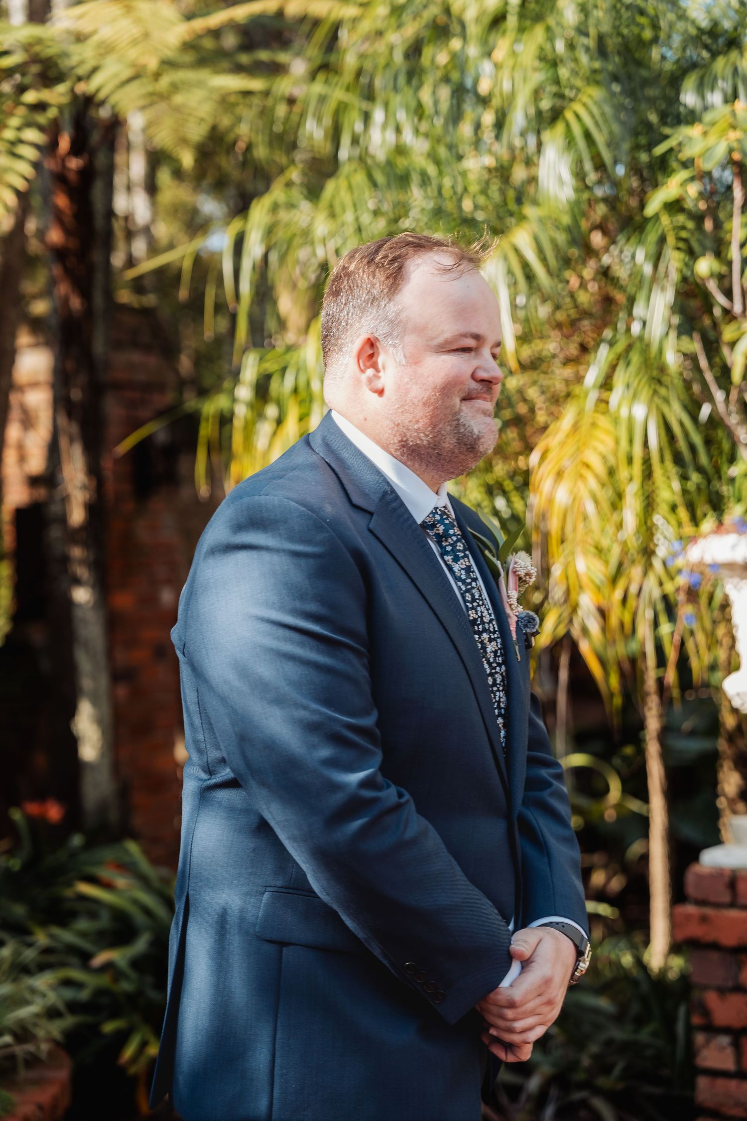 A groom in a navy suit stands thoughtfully in front of tropical palm trees during an outdoor wedding.