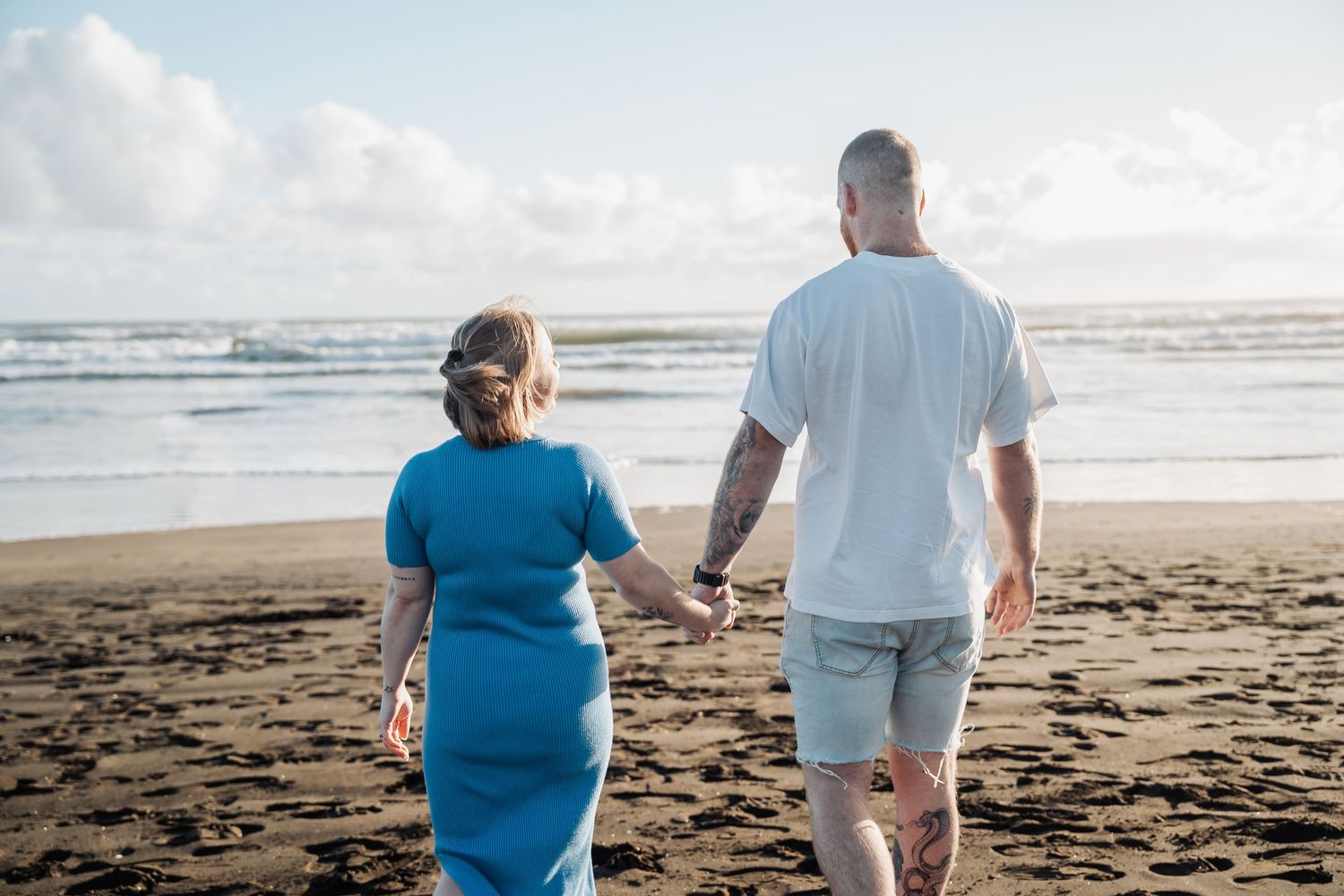 A couple holds hands while walking along a sandy beach at sunset with ocean waves in the background.