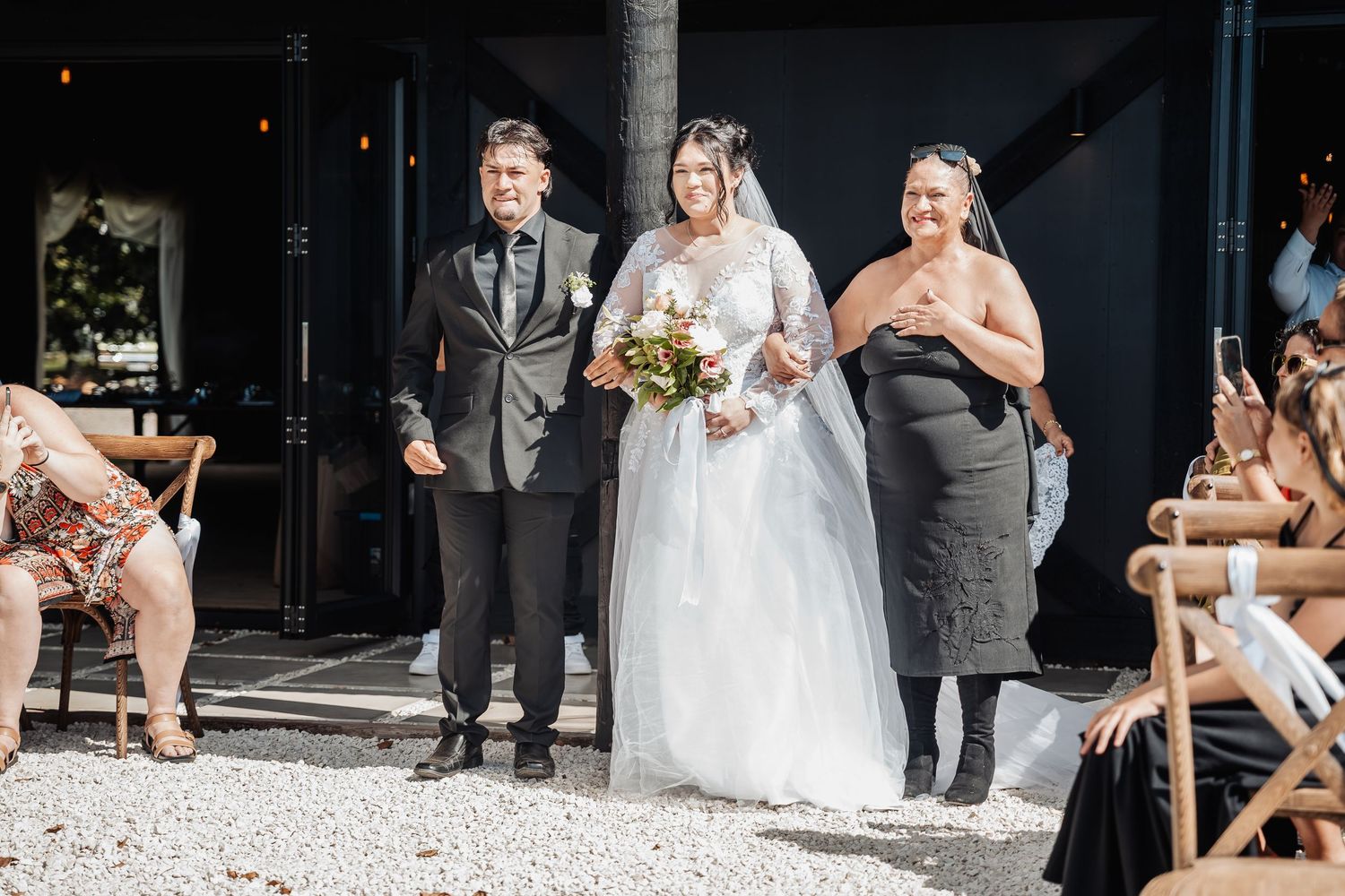 A bride in a white lace dress walks down an outdoor aisle with sunflower bouquet accompanied by two attendants.
