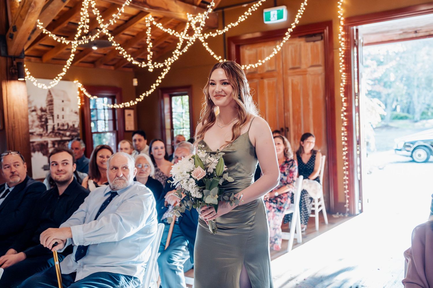 Beautiful wedding ceremony venue decorated with string lights as bridesmaid walks down rustic wooden aisle.
