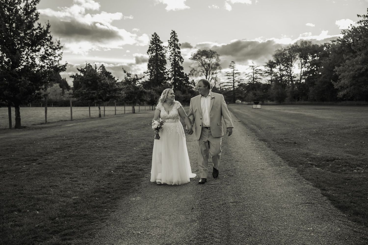 Black and white photo of bride and groom walking hand in hand down tree-lined path.