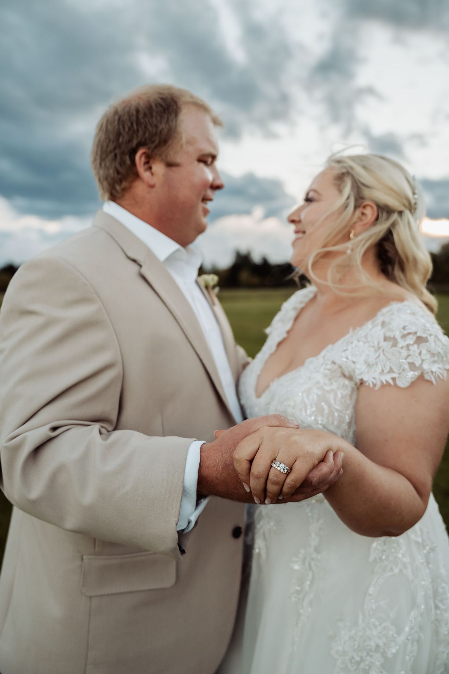 Bride and groom holding hands showing wedding ring with dramatic cloudy sky backdrop outdoors.