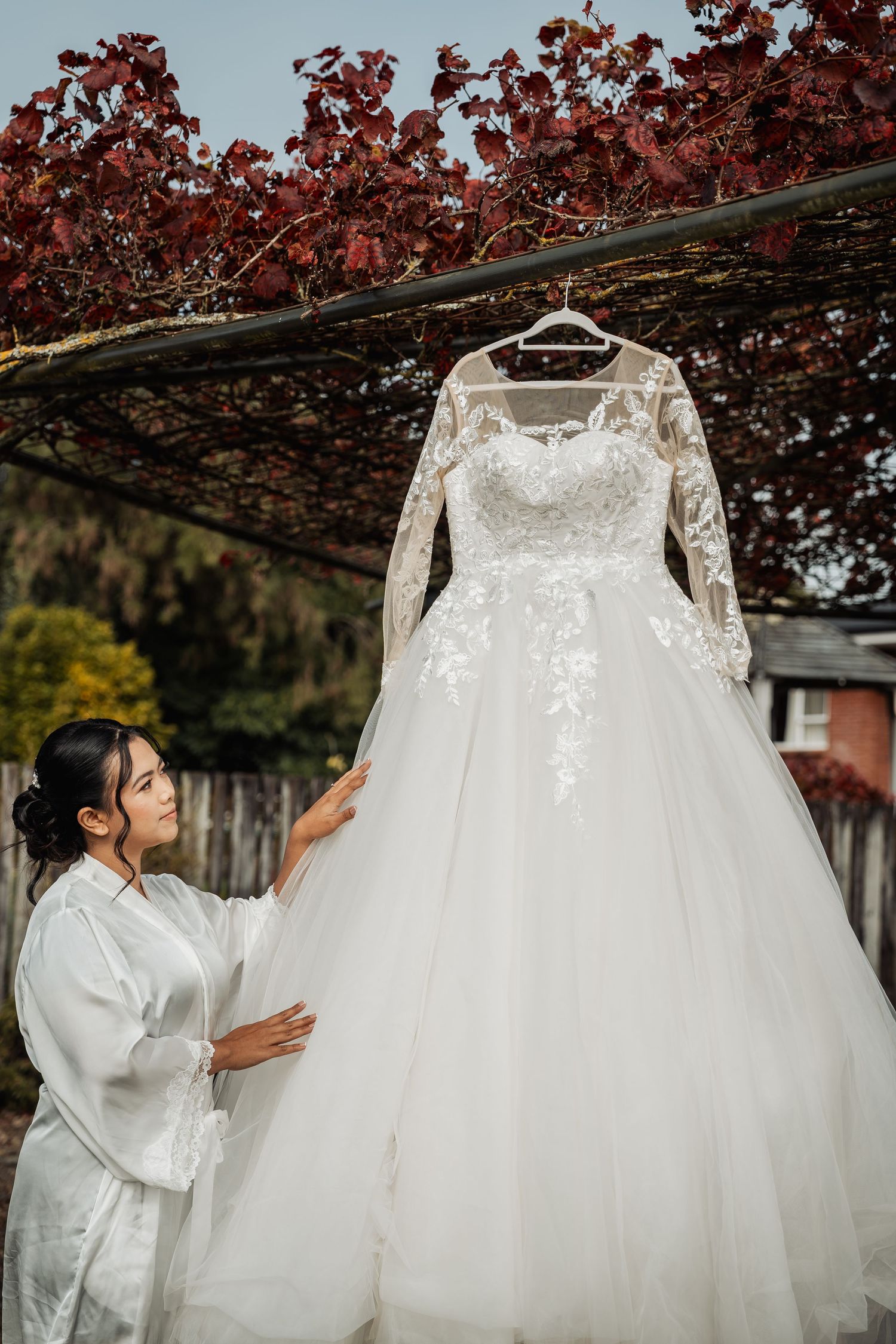 Bride in silk robe touching her elegant lace long-sleeve wedding dress hanging in a garden pergola.