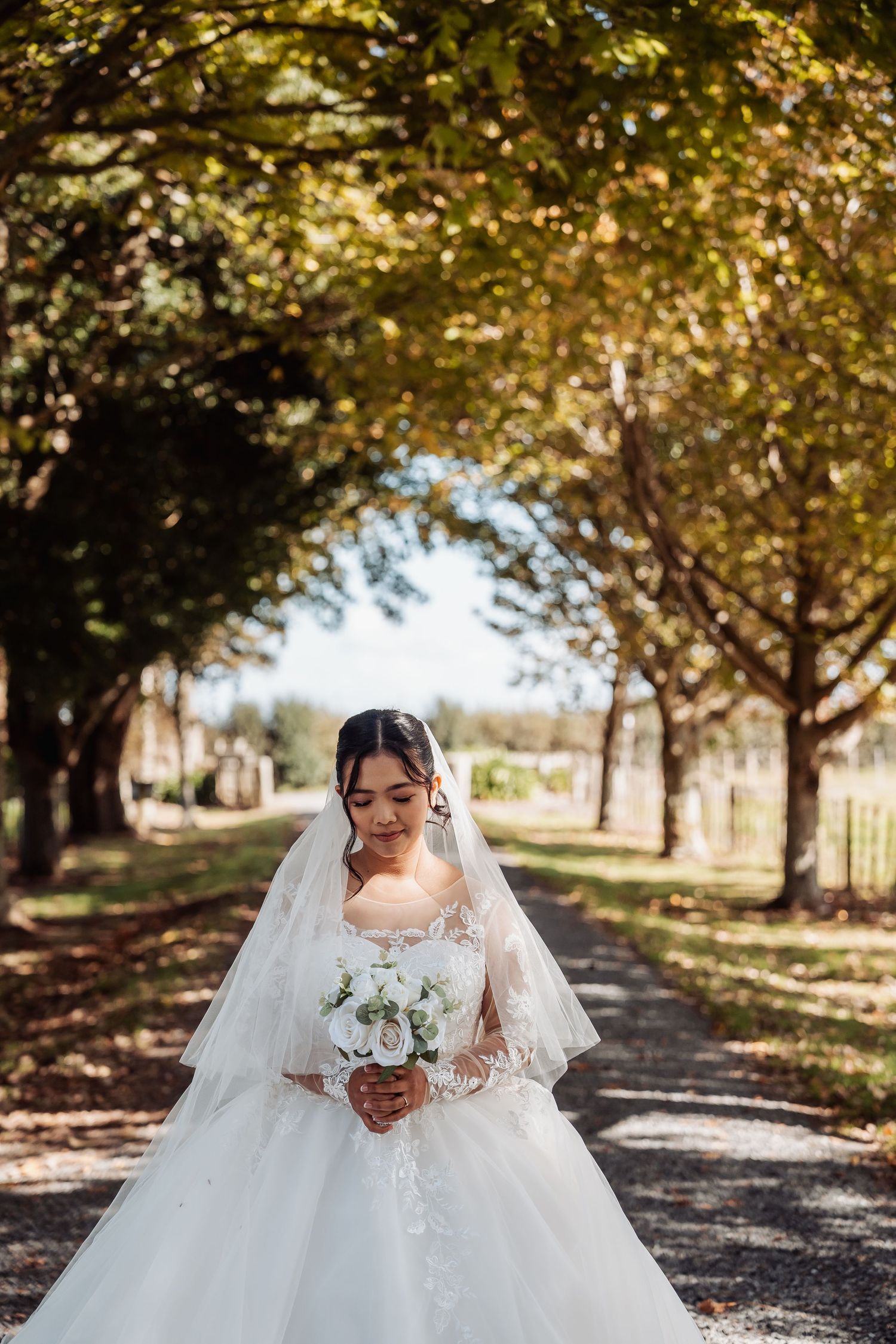 Bride in lace wedding gown looks down at white bouquet on sunlit tree-lined pathway.
