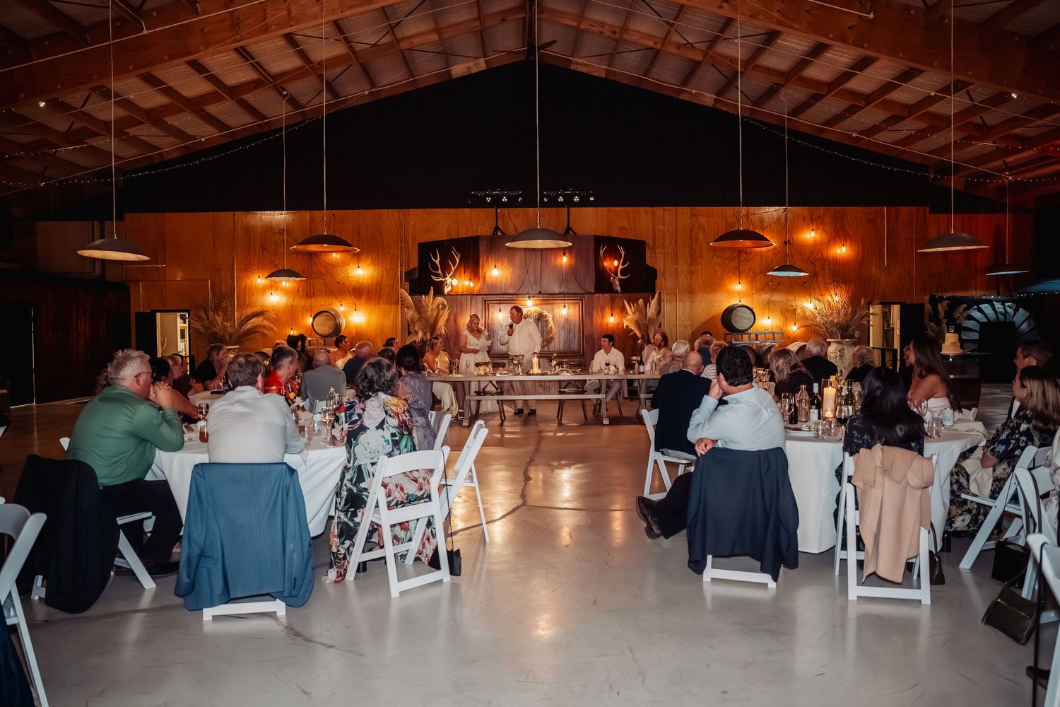 Guests seated at round tables in a rustic barn wedding reception hall with warm lighting and a stage backdrop.