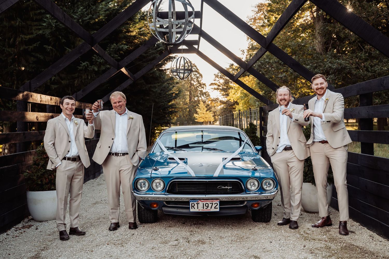 Four groomsmen in tan suits posing with classic blue vintage Mustang at elegant outdoor wedding venue.