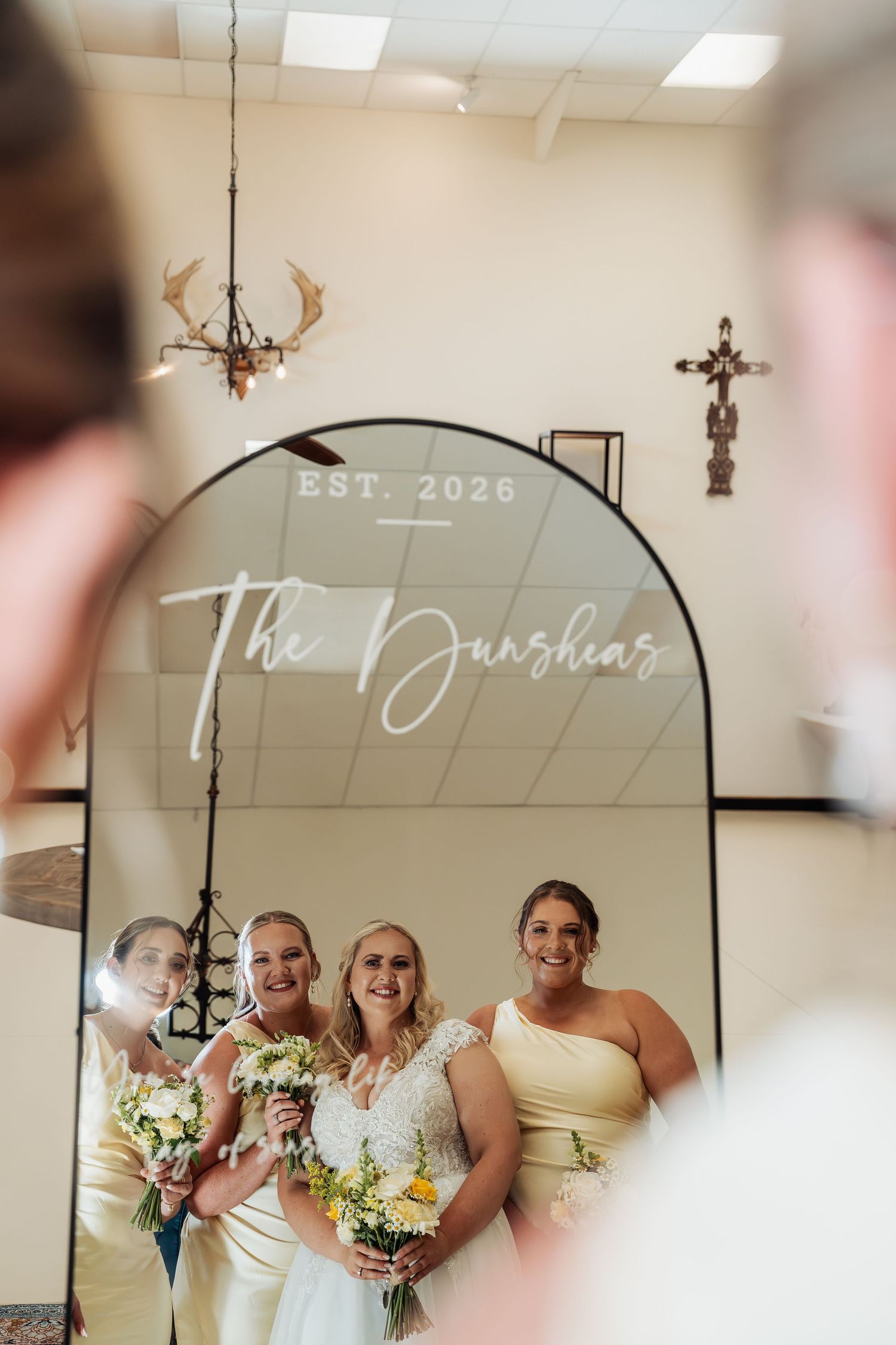 Bride and bridesmaids smiling in front of an arched mirror reading The Dunshias Est. 2026 holding yellow bouquets.