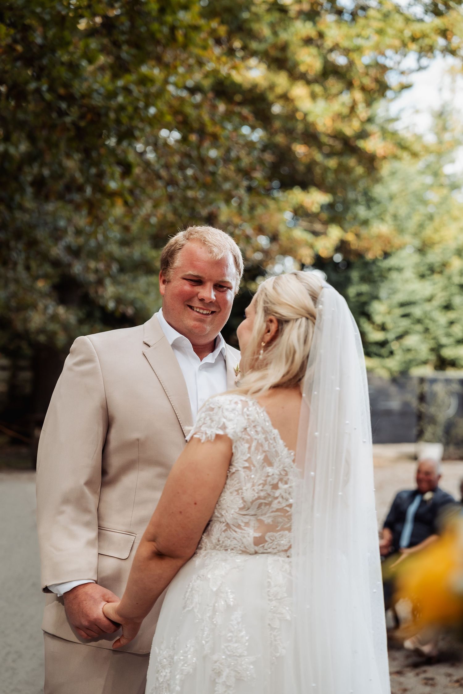 Smiling groom in tan suit holds hands with bride in lace wedding gown and veil outdoors.