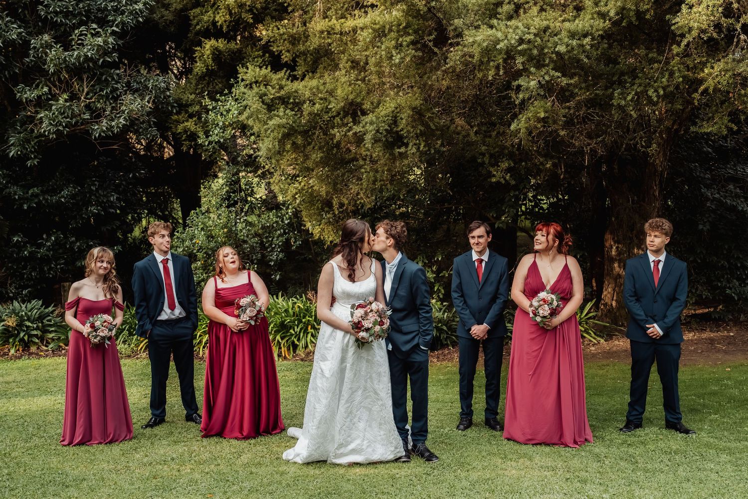 Wedding party in burgundy and navy poses on lawn with bride and groom kissing at center.