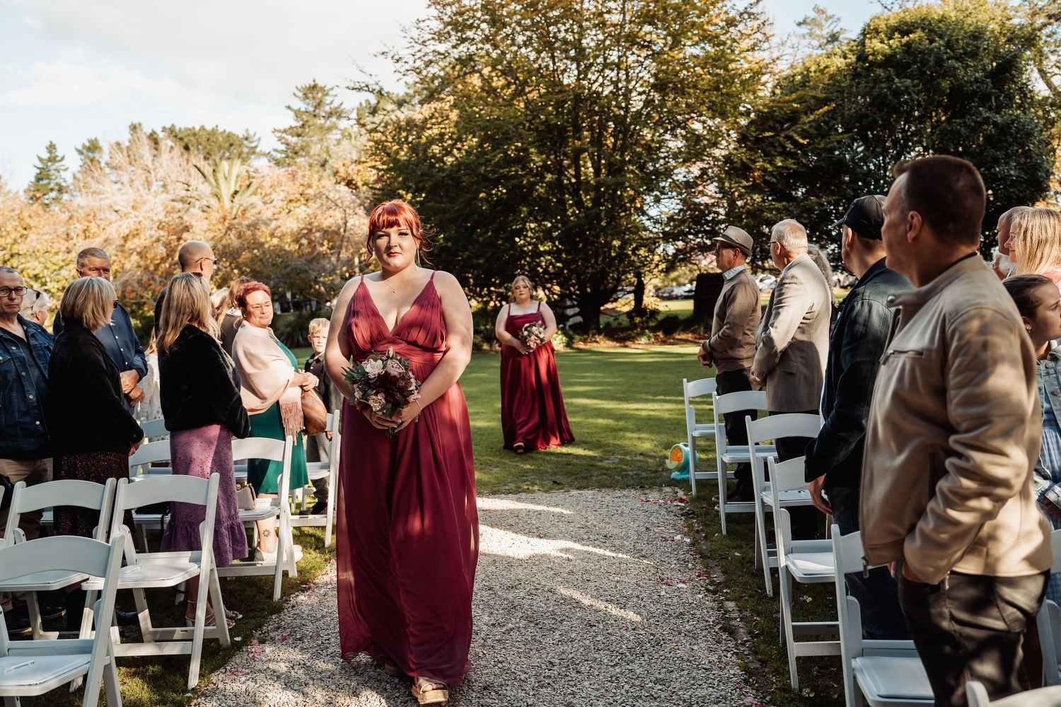 Bridesmaid in burgundy gown walks down outdoor wedding aisle lined with white chairs and guests.