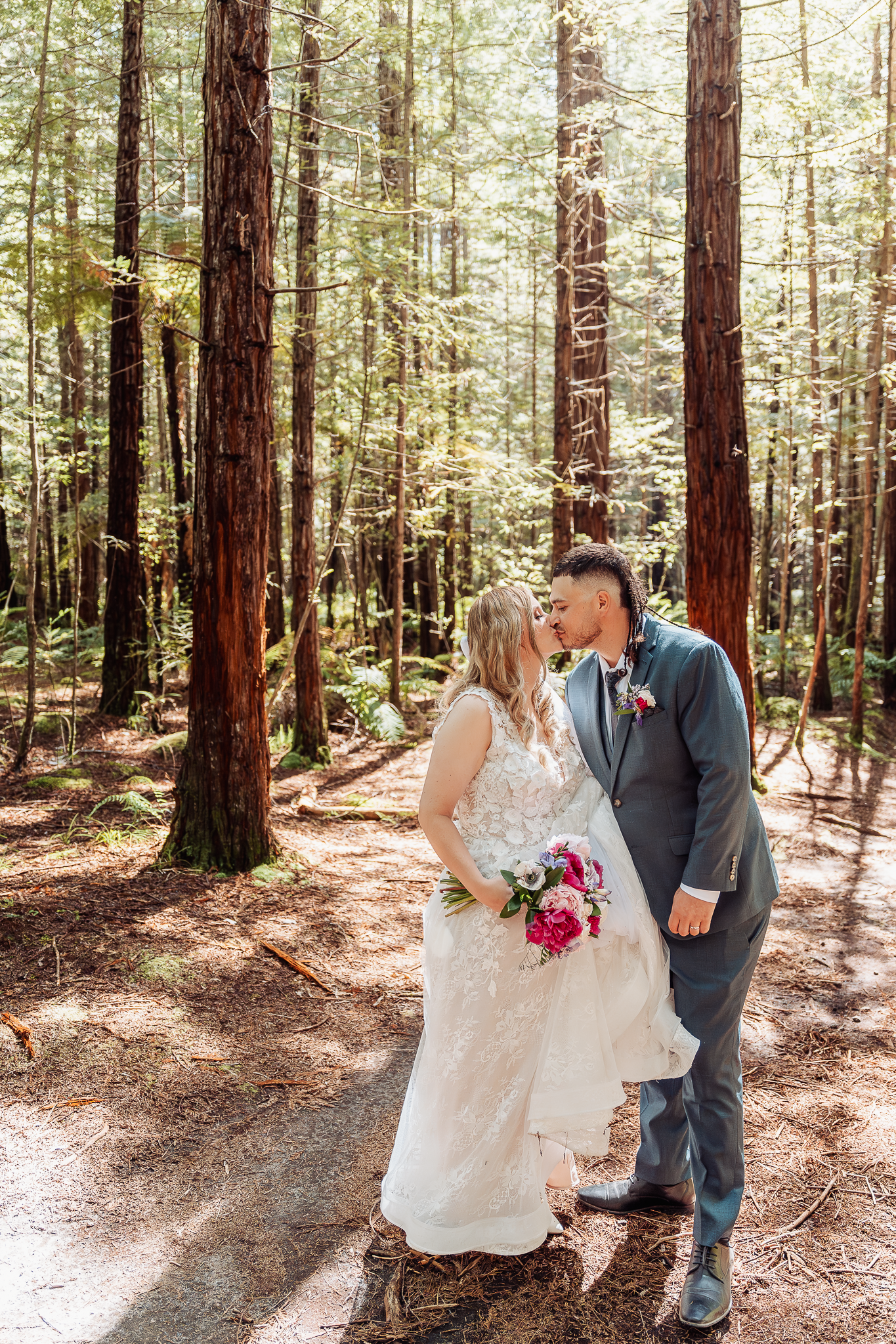 A romantic wedding photo captured in a sunlit forest with towering redwood trees in the background.