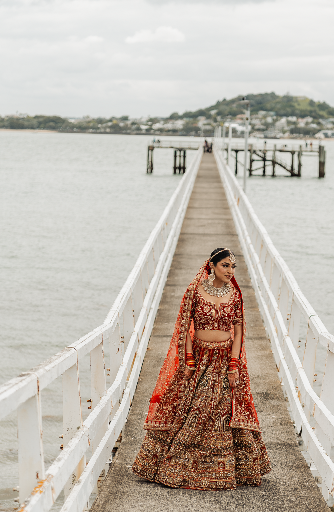 Traditional Indian bride in red lehenga photographed on a long white pier with water views.