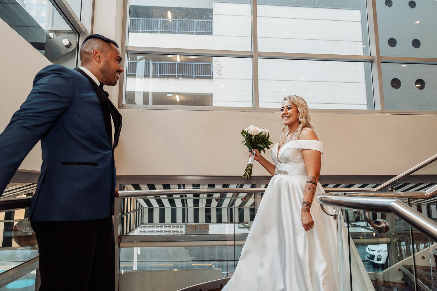 Wedding couple stands on modern staircase with large windows and natural lighting.