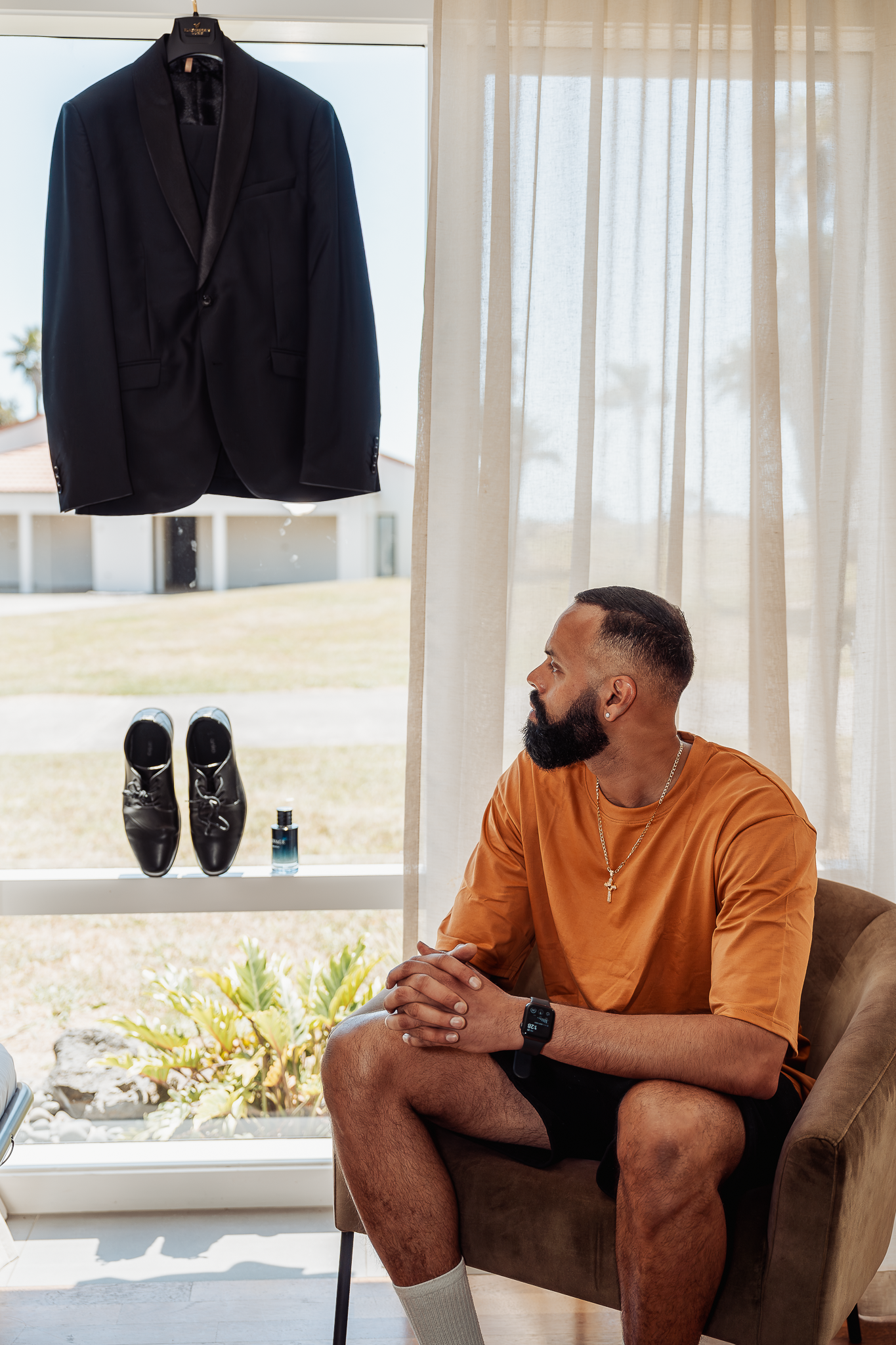 A formal black suit jacket and dress shoes displayed near sheer curtains in a bright room.