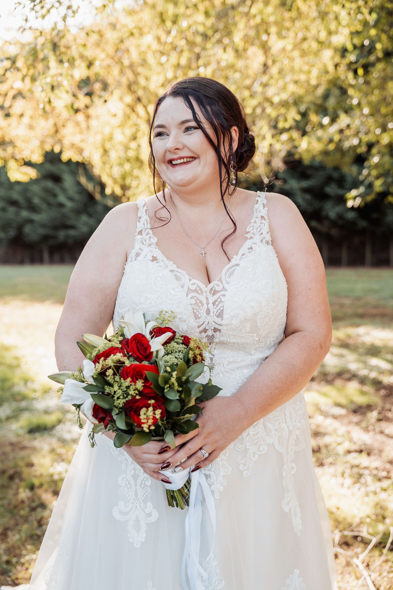 Radiant bride in lace wedding dress holds red and white bouquet in sunlit garden setting.