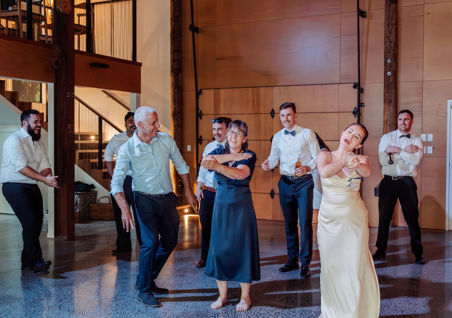 Wedding party dancing energetically in a grand venue with wooden staircase and warm lighting.