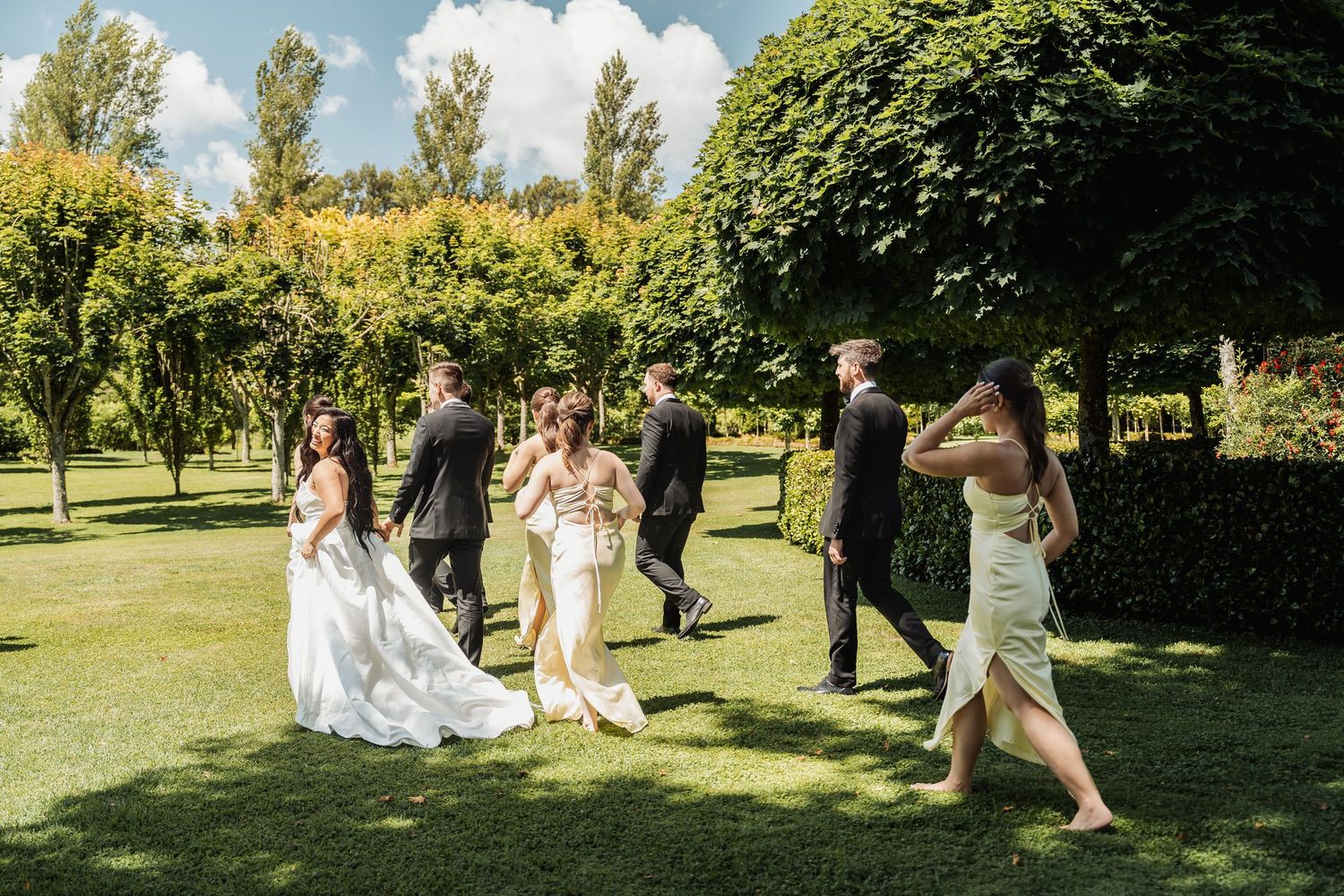 Wedding party walks through lush green garden grounds on a sunny day.