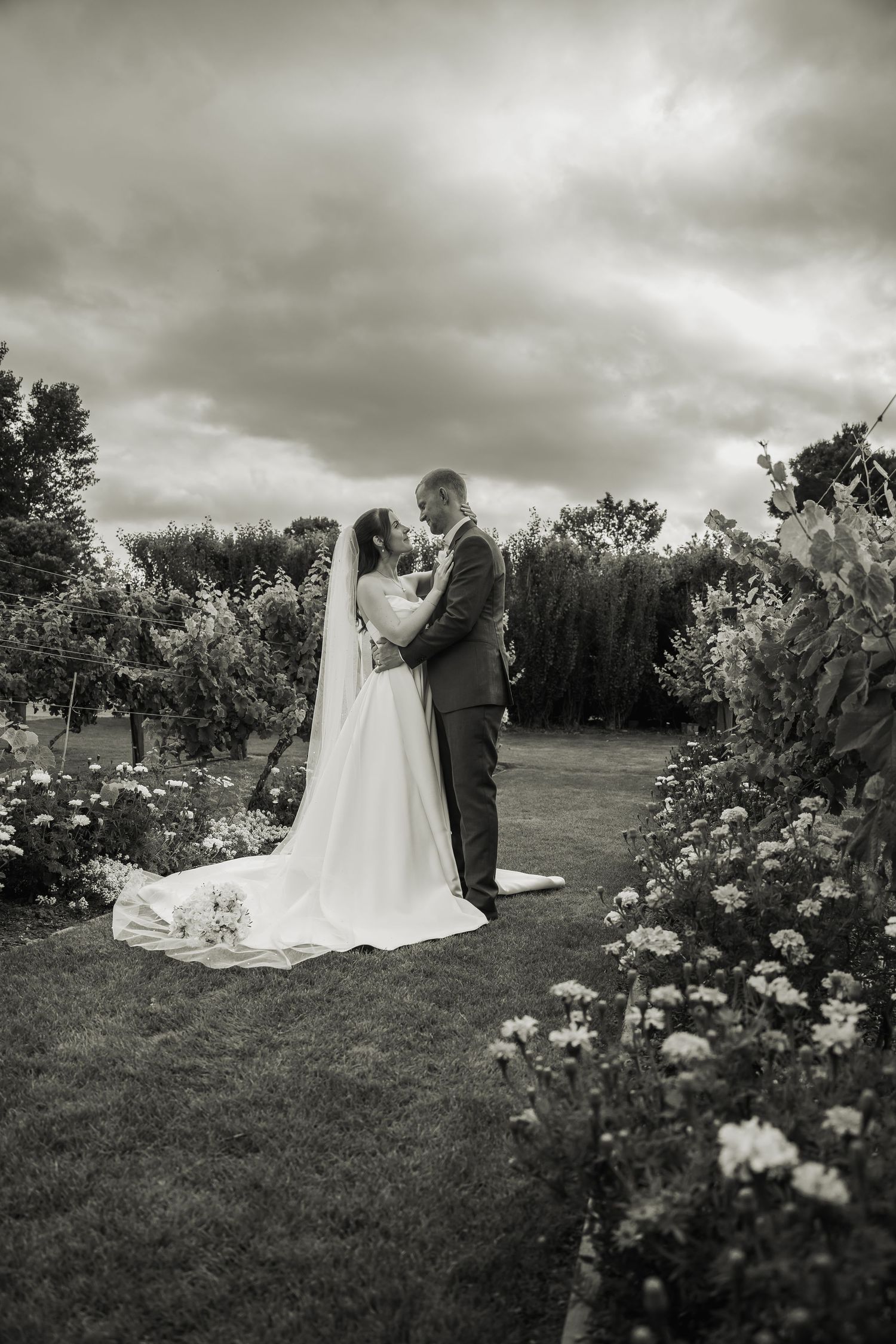 Black and white portrait of bride and groom embracing in flower-lined garden path under dramatic cloudy sky.