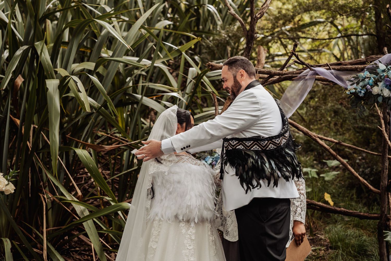 Groom in black feather vest adjusts brides white veil during outdoor forest wedding ceremony.