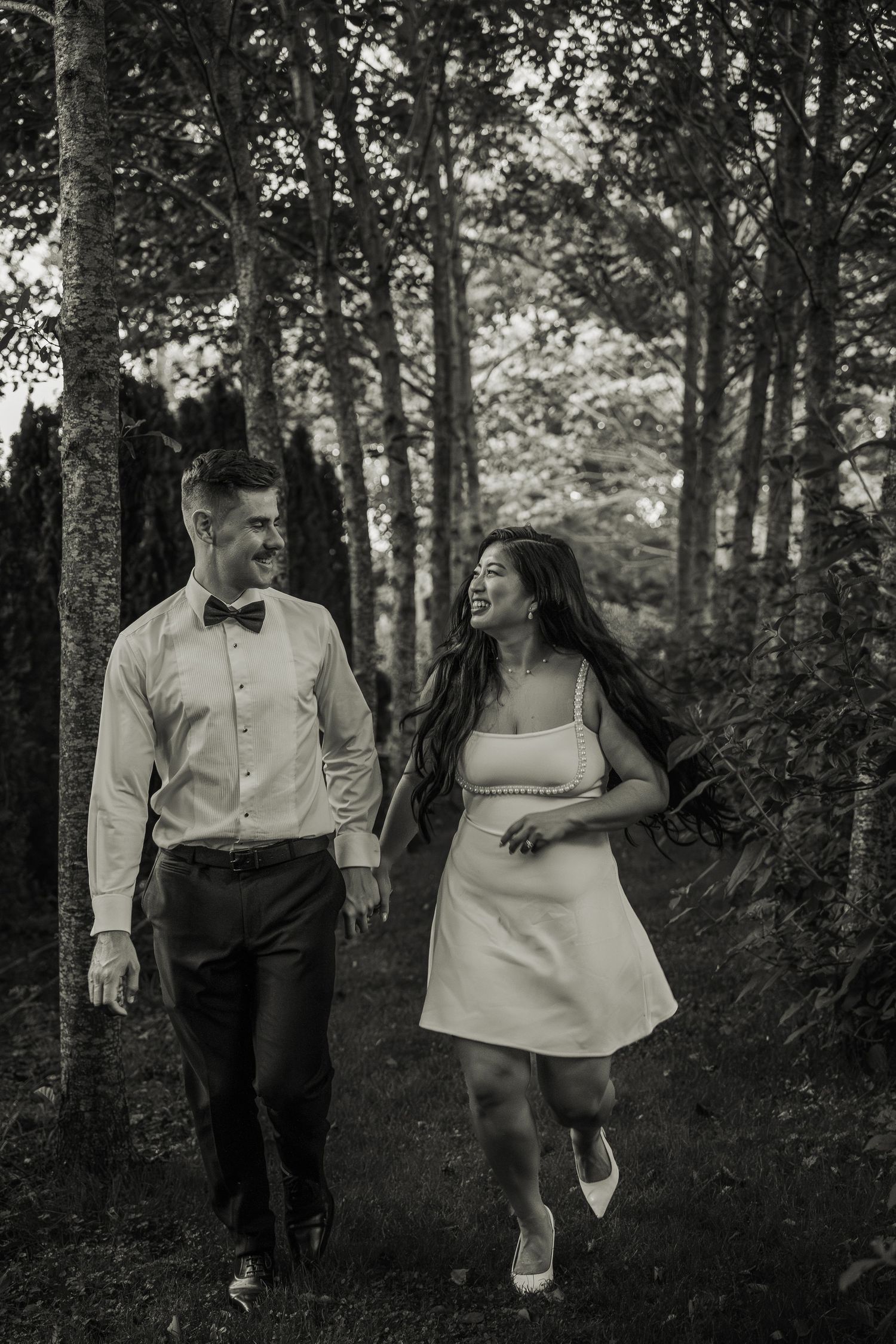 Black and white photo of smiling bride and groom walking together through wooded garden path.
