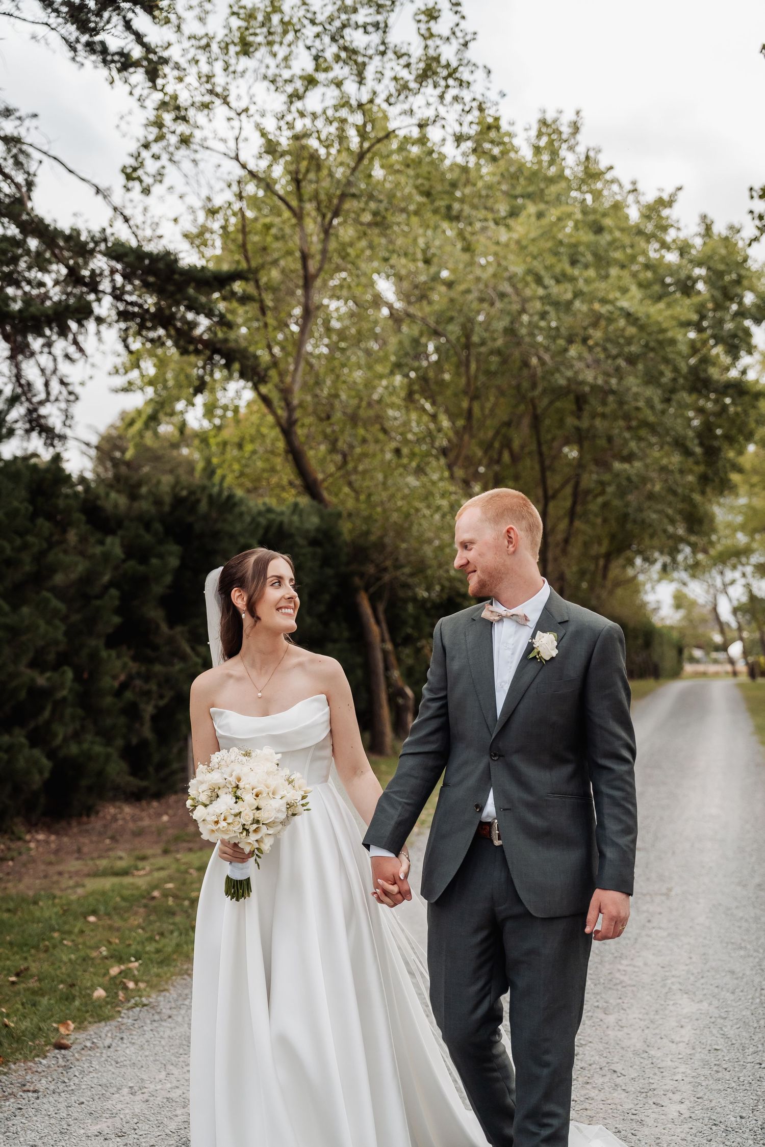 Smiling bride and groom holding hands while walking down a tree-lined gravel path on their wedding day.