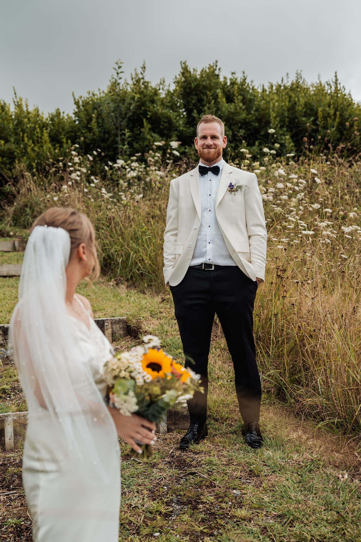 Groom in white tuxedo smiling at bride holding sunflower bouquet during outdoor first look.